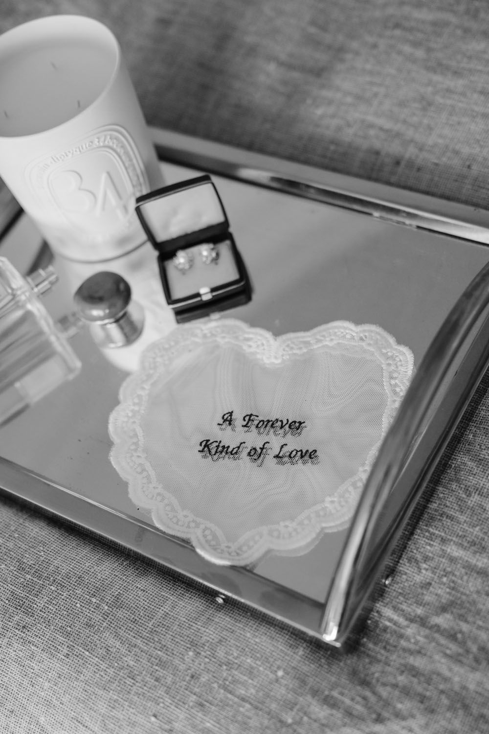 Black and white photo of a mirrored tray with wedding jewelry, perfume, and a heart lace handkerchief reading A Forever Kind of Love.
