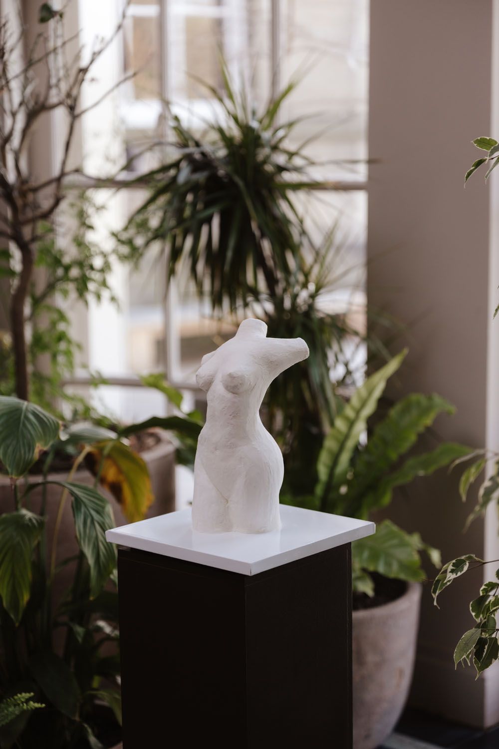 White marble female torso sculpture displayed on black pedestal surrounded by lush green tropical plants near window.