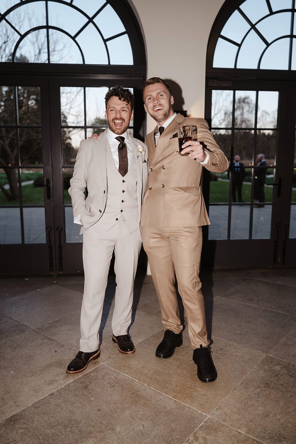 Two men in light-colored suits pose together in front of arched windows in an elegant indoor setting.