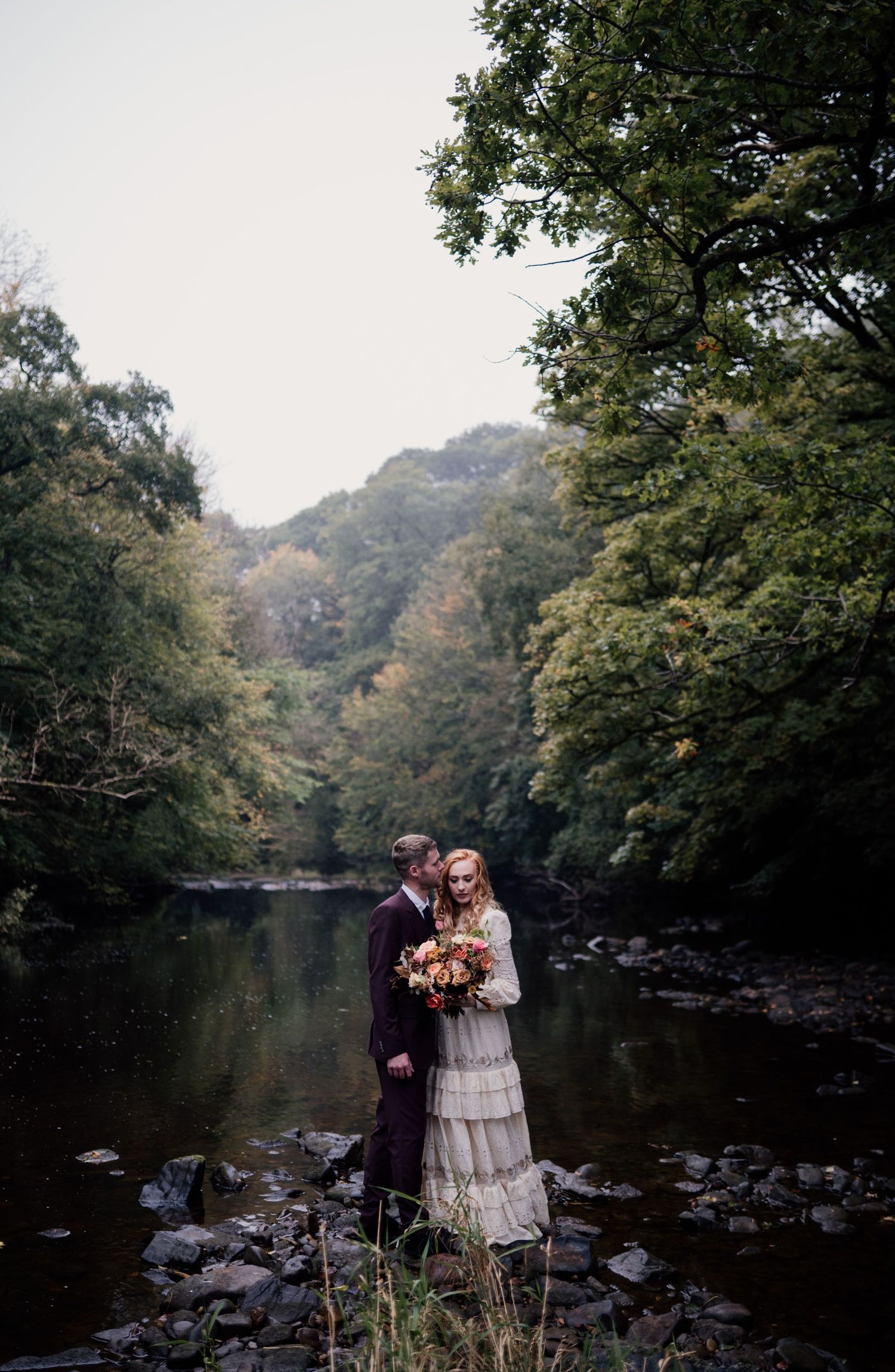 Couple smiling on their wedding day, stood on rocks on a river with trees surrounding them.