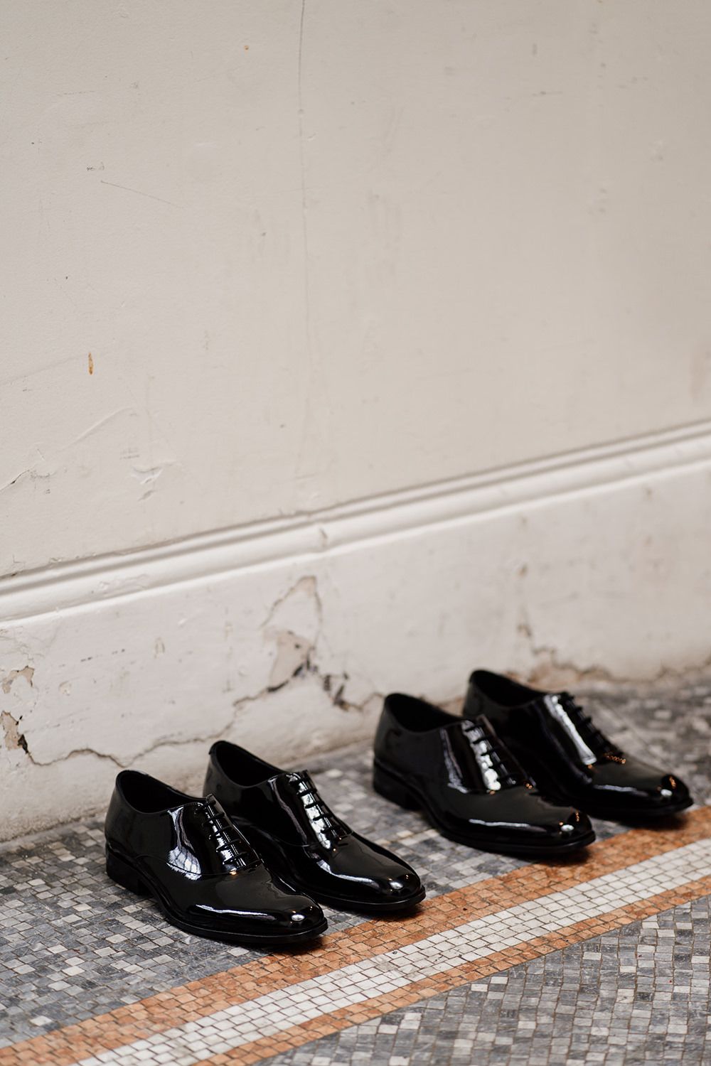Two pairs of black shiny wedding shoes on mosaic flooring with white walls behind.