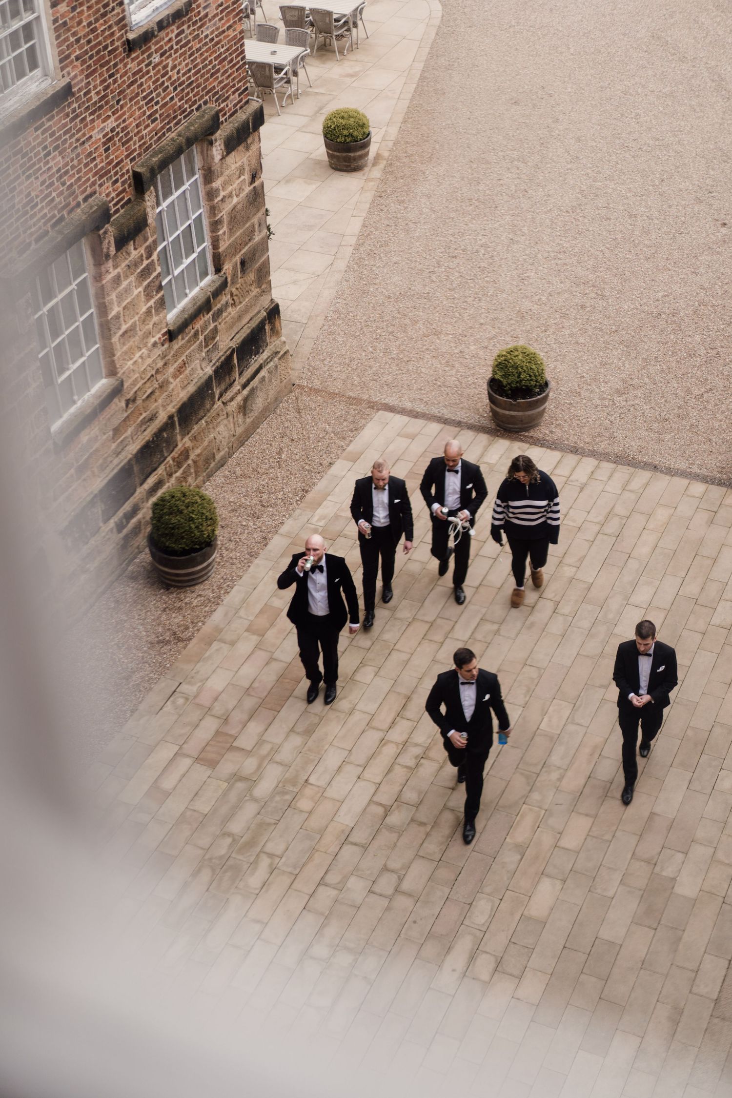 Men in black suits walking across a courtyard surrounded by brick walls and decorative potted plants.