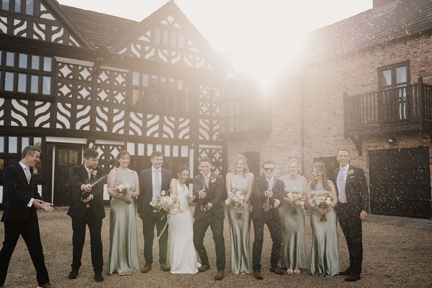 Bride and Groom standing with a bottle of champagne spraying surrounded by their wedding party with Holford Hall behind.