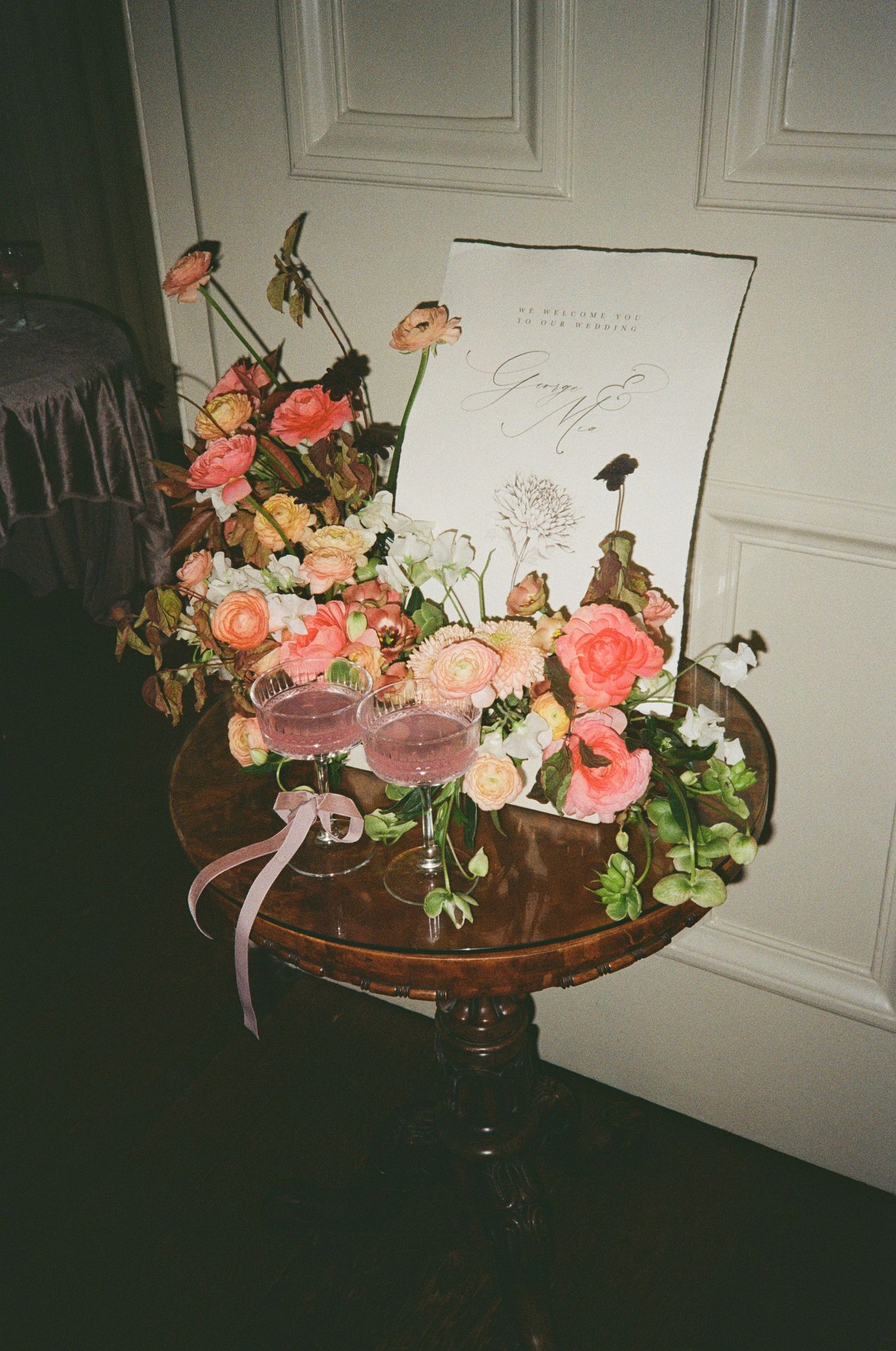 Romantic floral arrangement with pink roses and two champagne glasses on a vintage wooden table with a wedding sign.