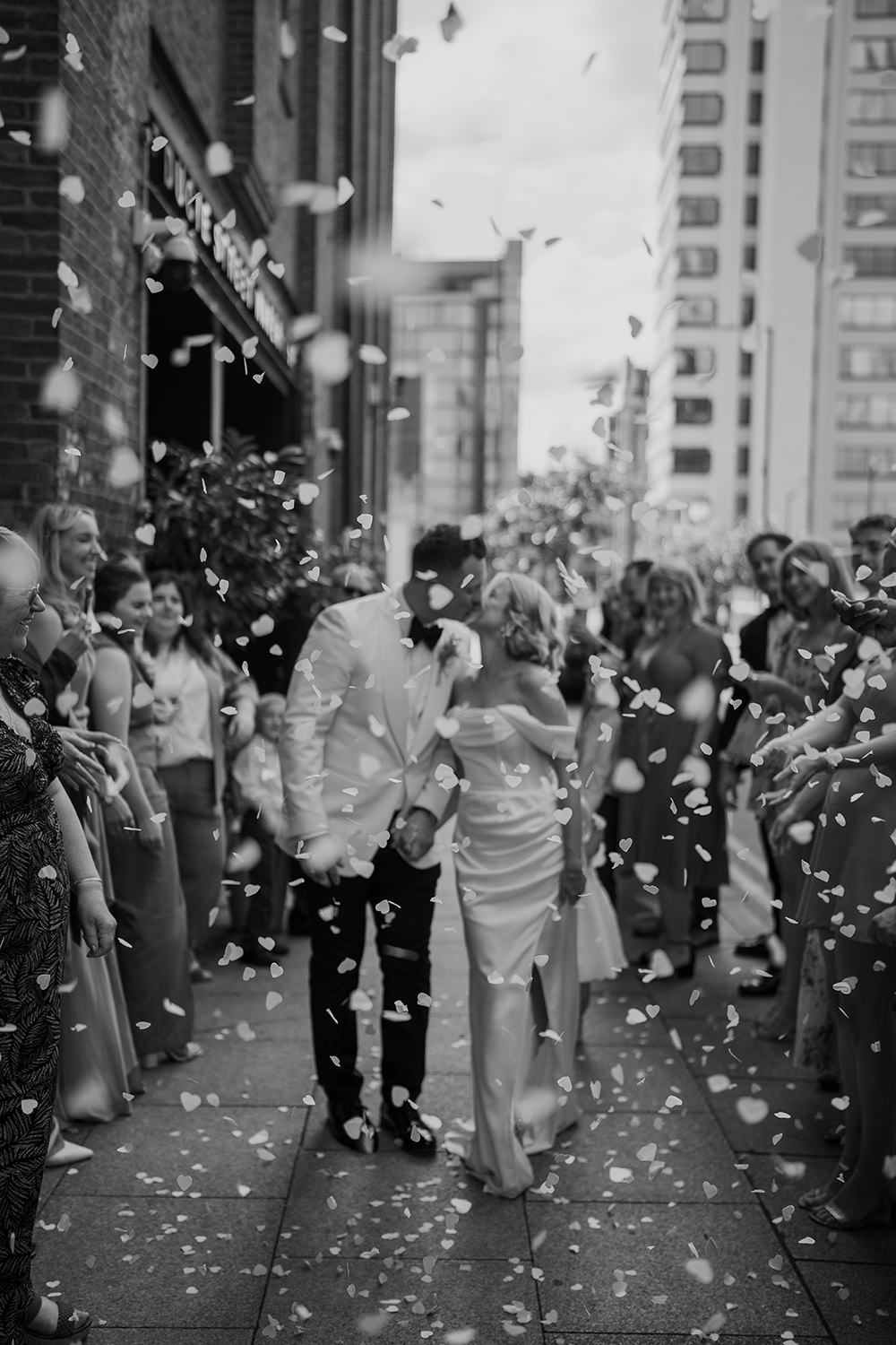 A joyful wedding exit with guests throwing white rose petals as the couple walks down the steps in black and white.