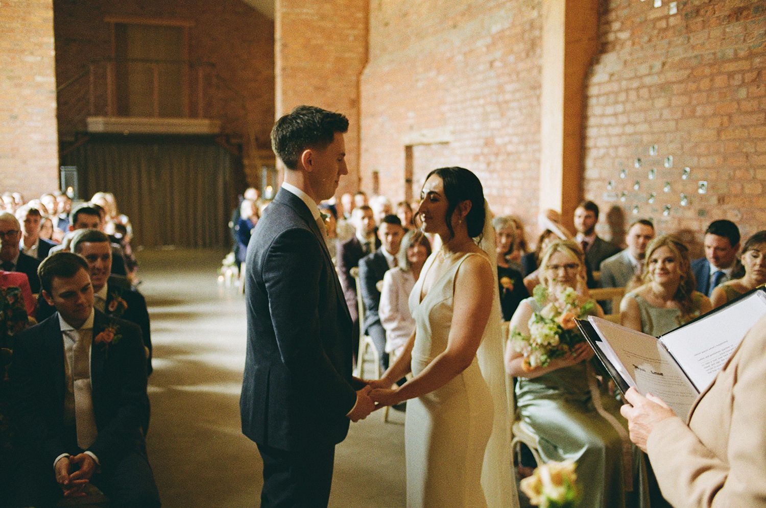 35mm Film image of bride and groom stood holding hands and smiling with wedding guests in the background.