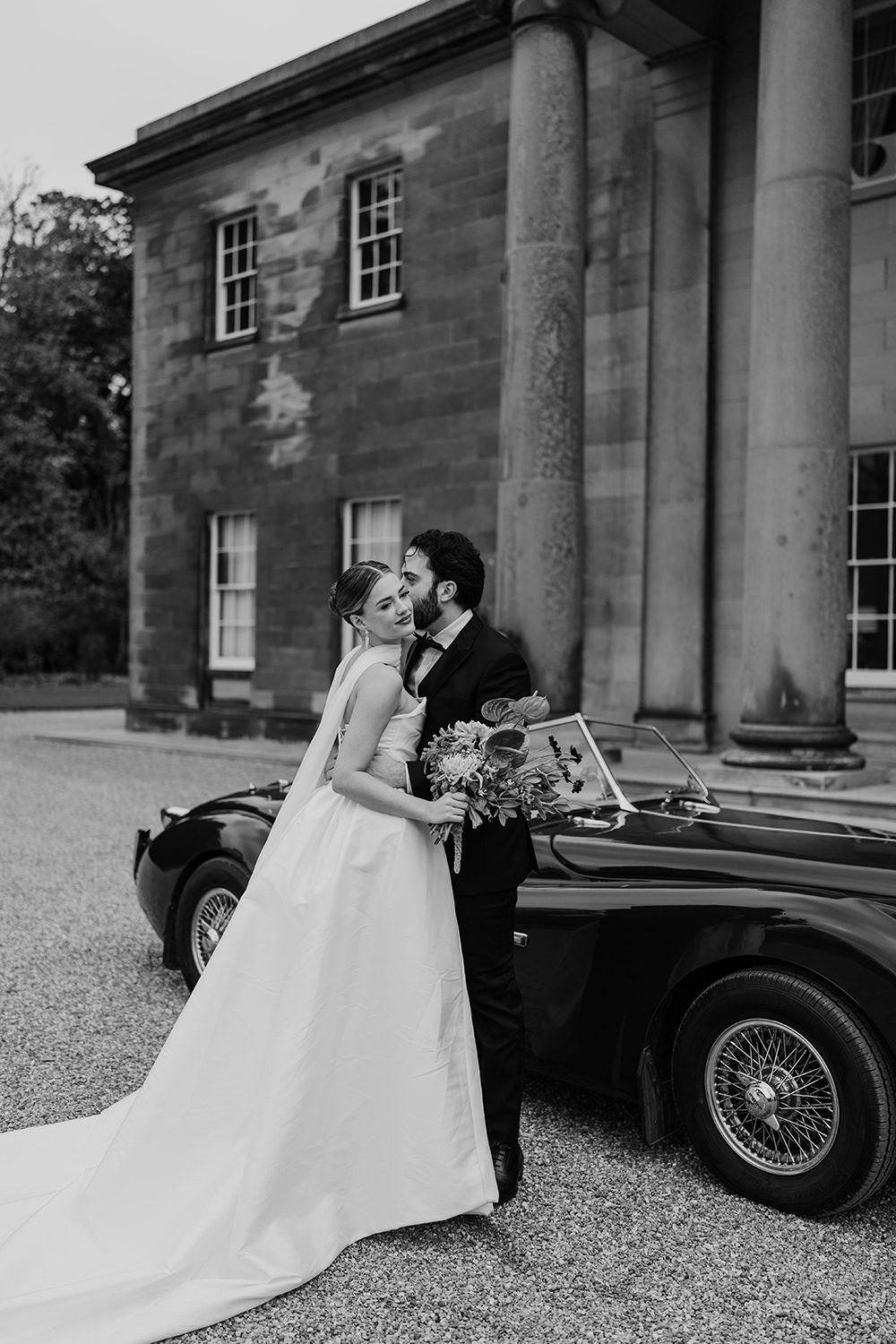 Bride and groom laughing and running through the grounds at Rise Hall with a vintage car in the background.