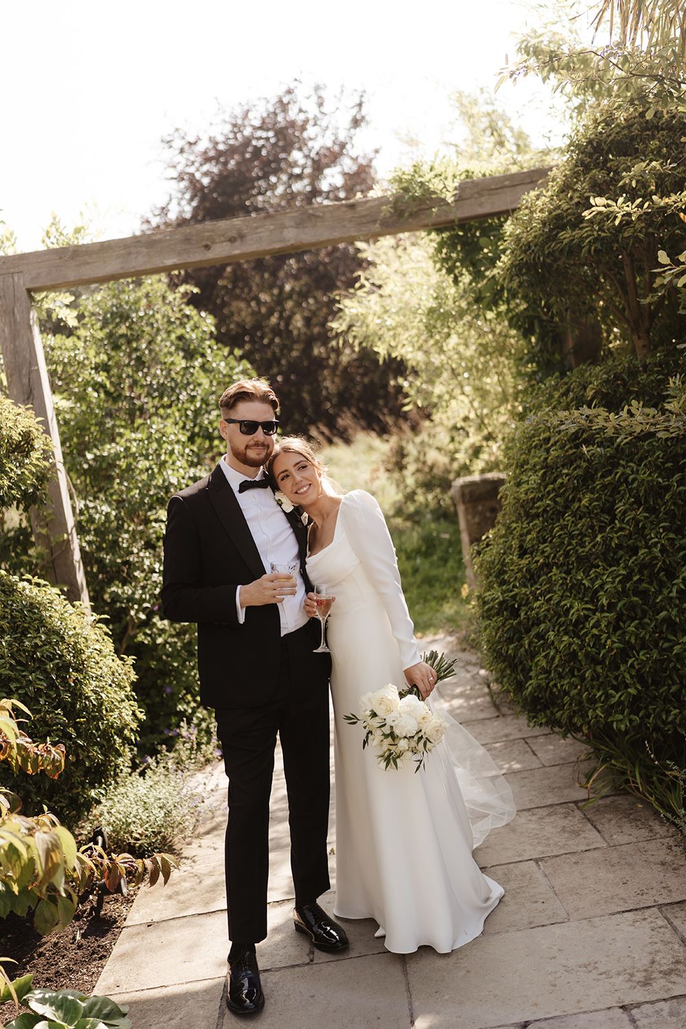 A couple in wedding attire shares intimate moments in a garden setting with stone pathways and lush greenery.