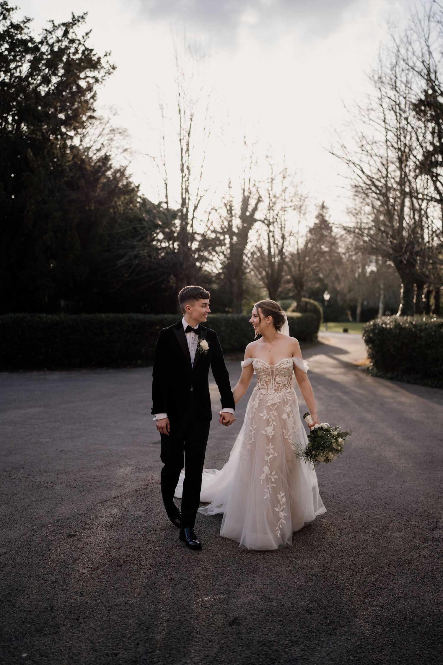 Bride and groom holding hands looking at each other as the sun sets amongst trees behind them.