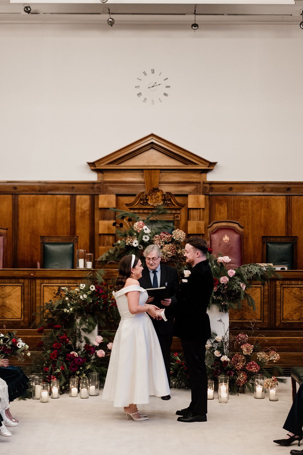 A wedding ceremony takes place in an ornate wood-paneled room with floral arrangements and candles decorating the altar area.