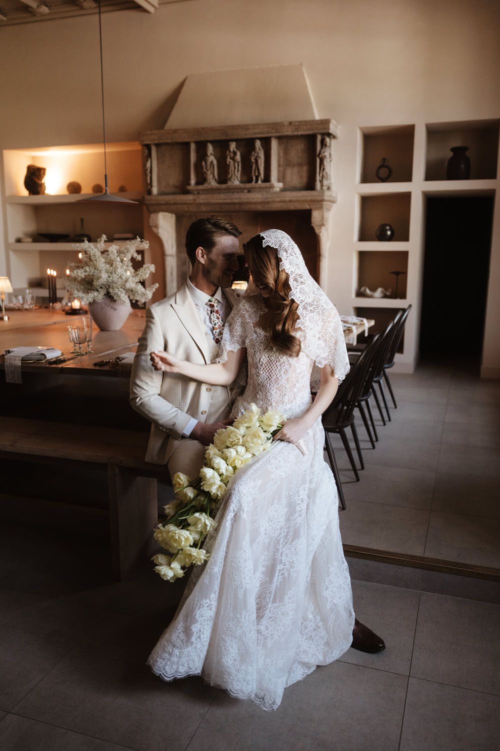 Bride and groom share a romantic kiss in a rustic wedding venue with stone fireplace, lace dress, and yellow floral bouquet.
