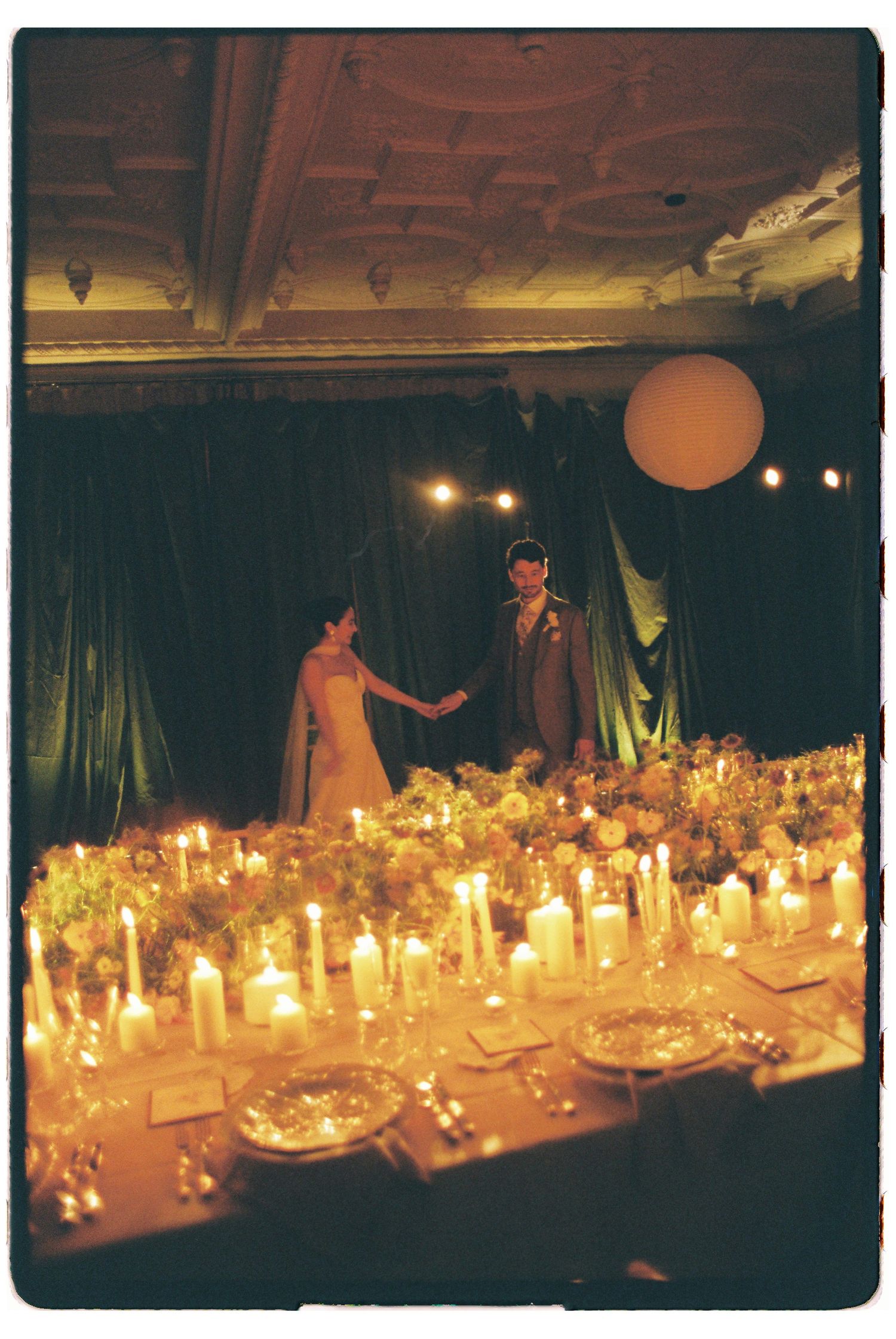 Romantic candlelit wedding reception with bride and groom holding hands on stage surrounded by floral arrangements and glowing candles.