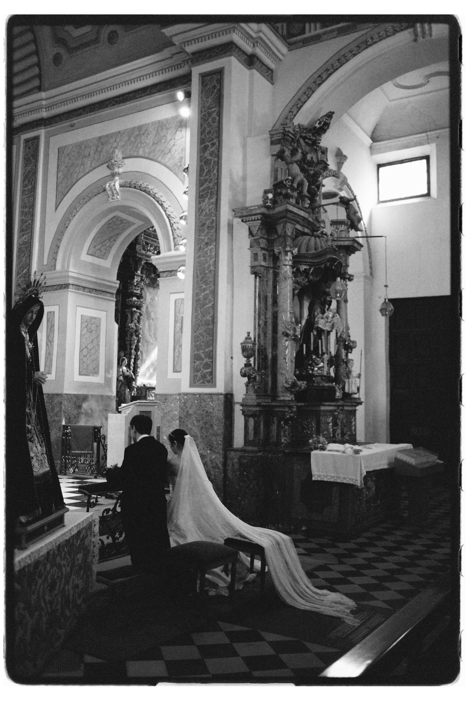 Black and white photo of bride and groom kneeling at the altar of an ornate baroque church during ceremony.
