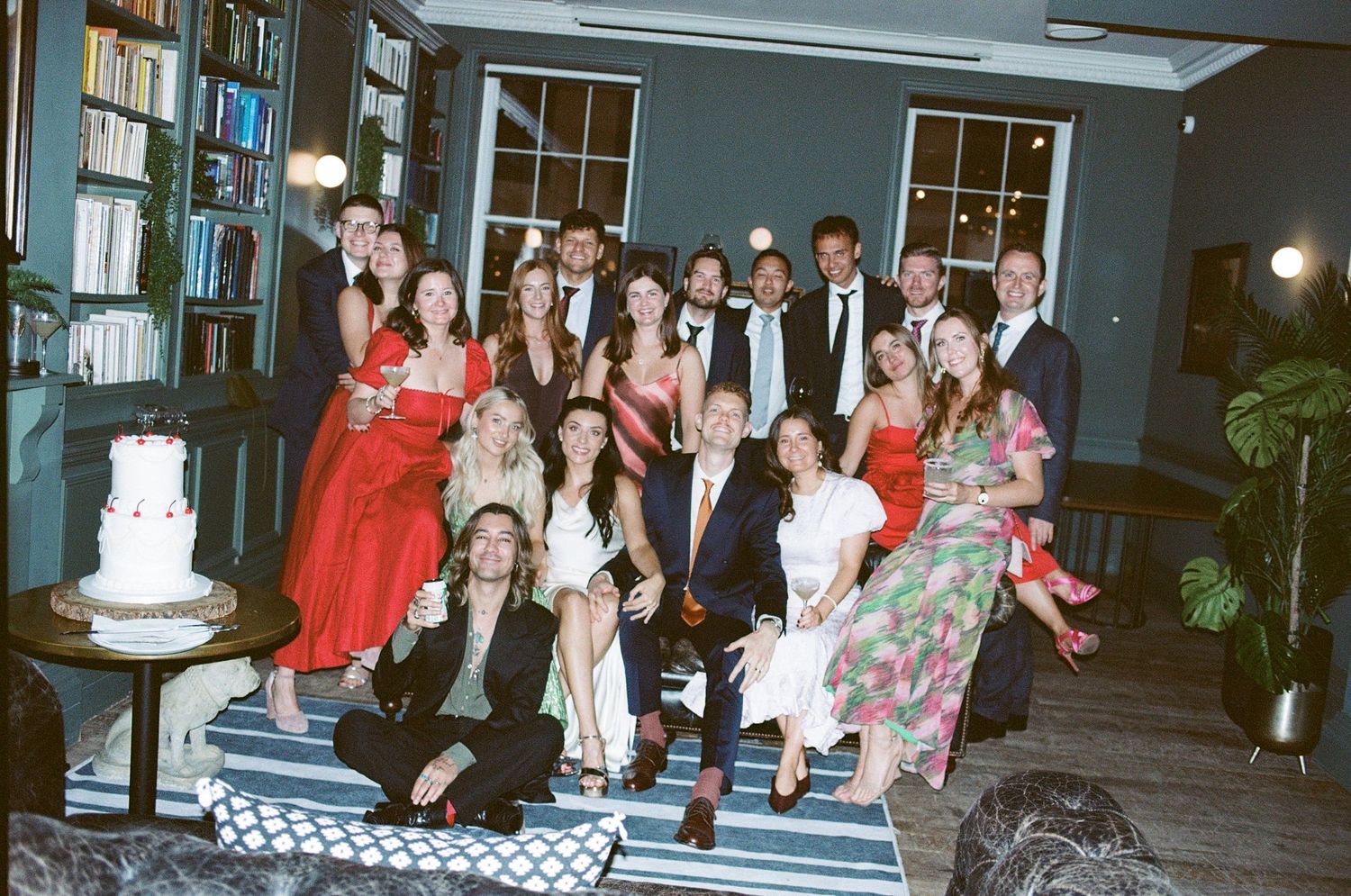 Large wedding party group photo taken indoors at a stylish venue with a white cake visible on the left.