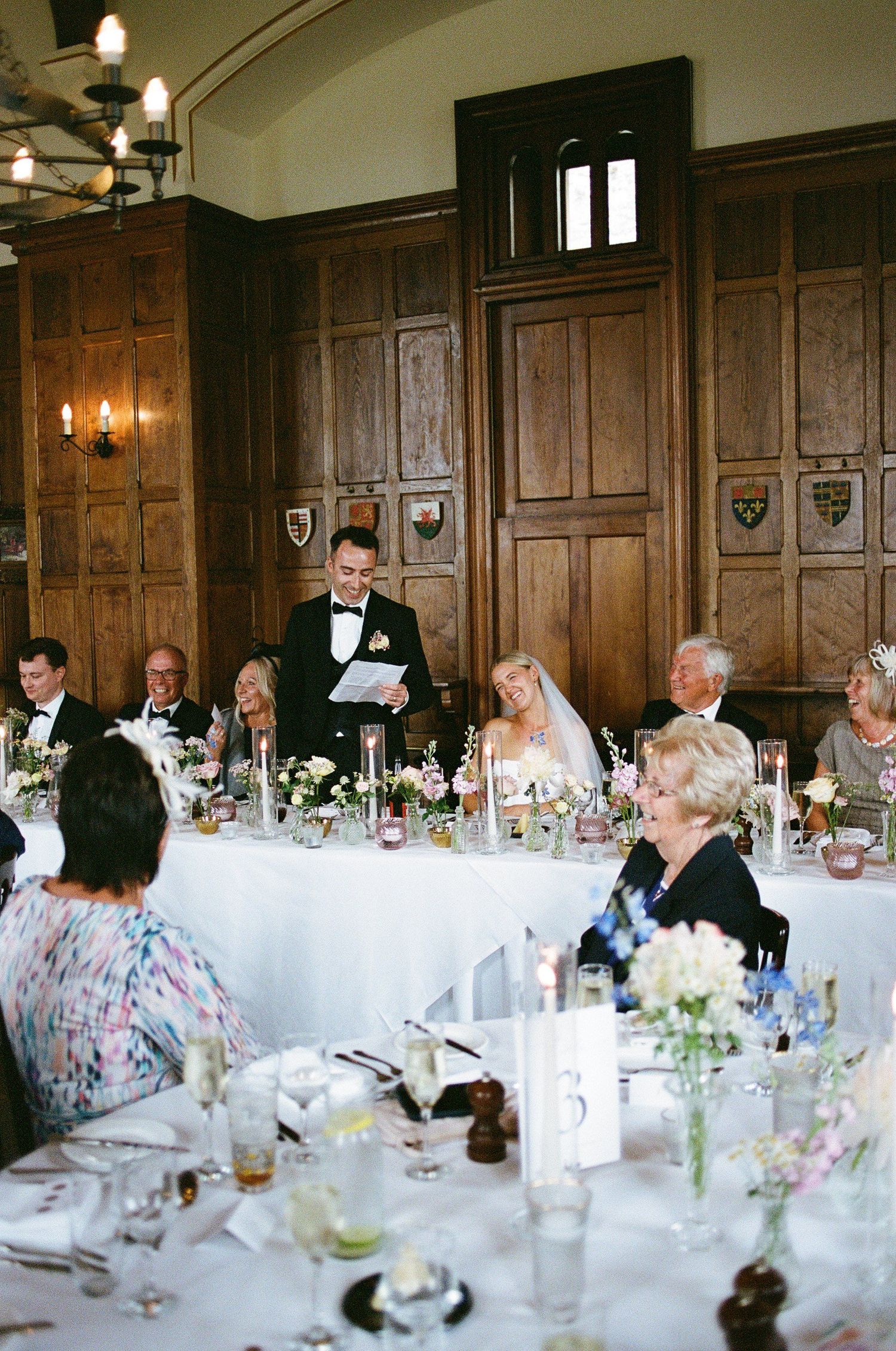 Groom delivers a speech at a wedding reception in an elegant wood-paneled dining hall.