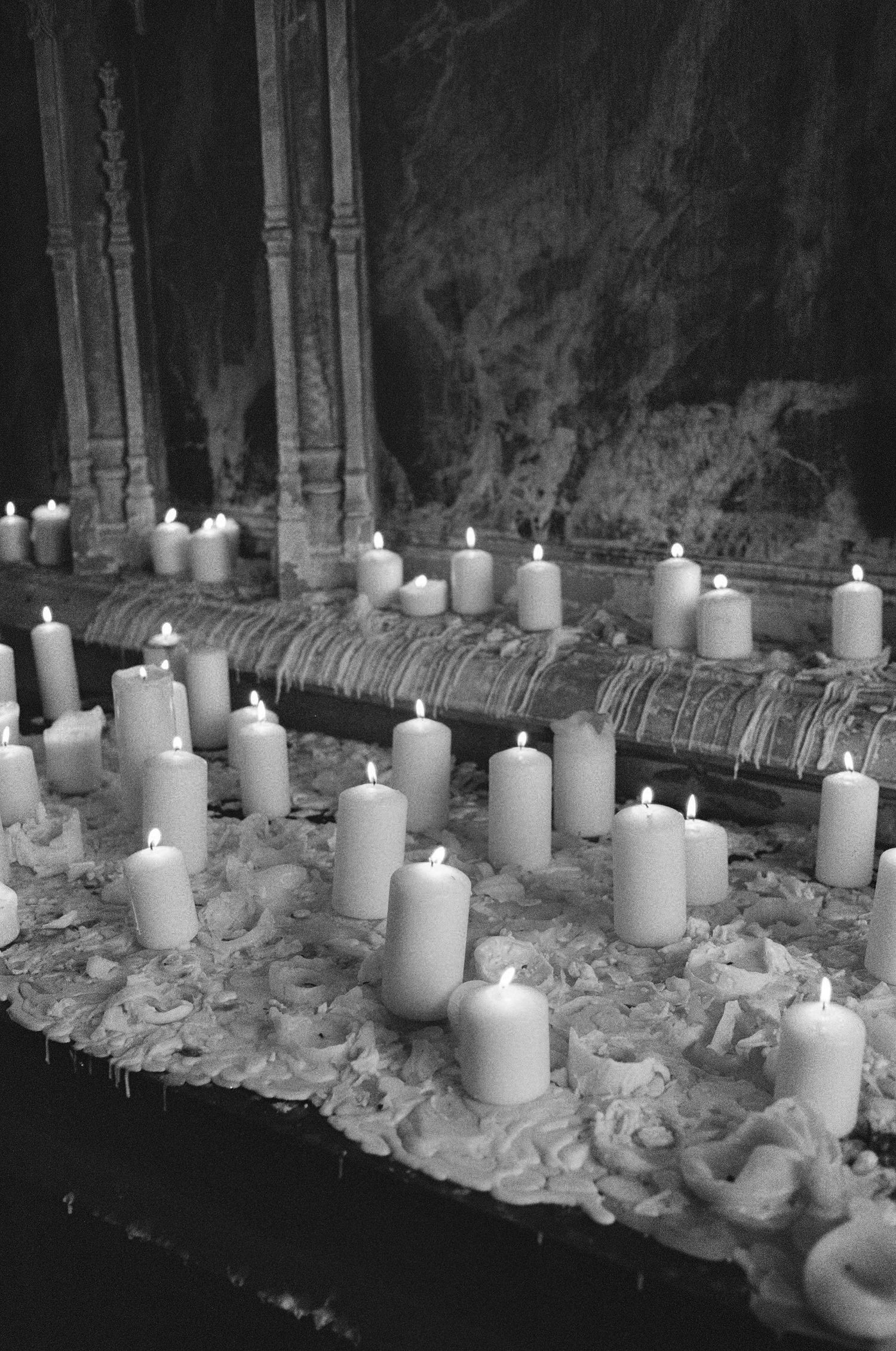 Black and white photo of multiple lit white pillar candles arranged on an ornate stone altar.