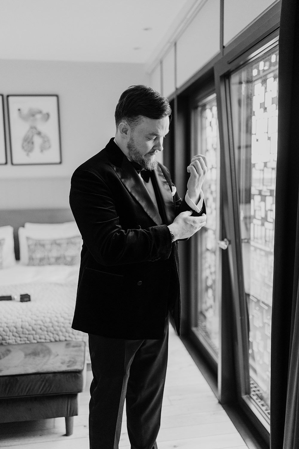 Black and white photo of person in velvet suit jacket adjusting cufflinks while standing near large windows and bed.