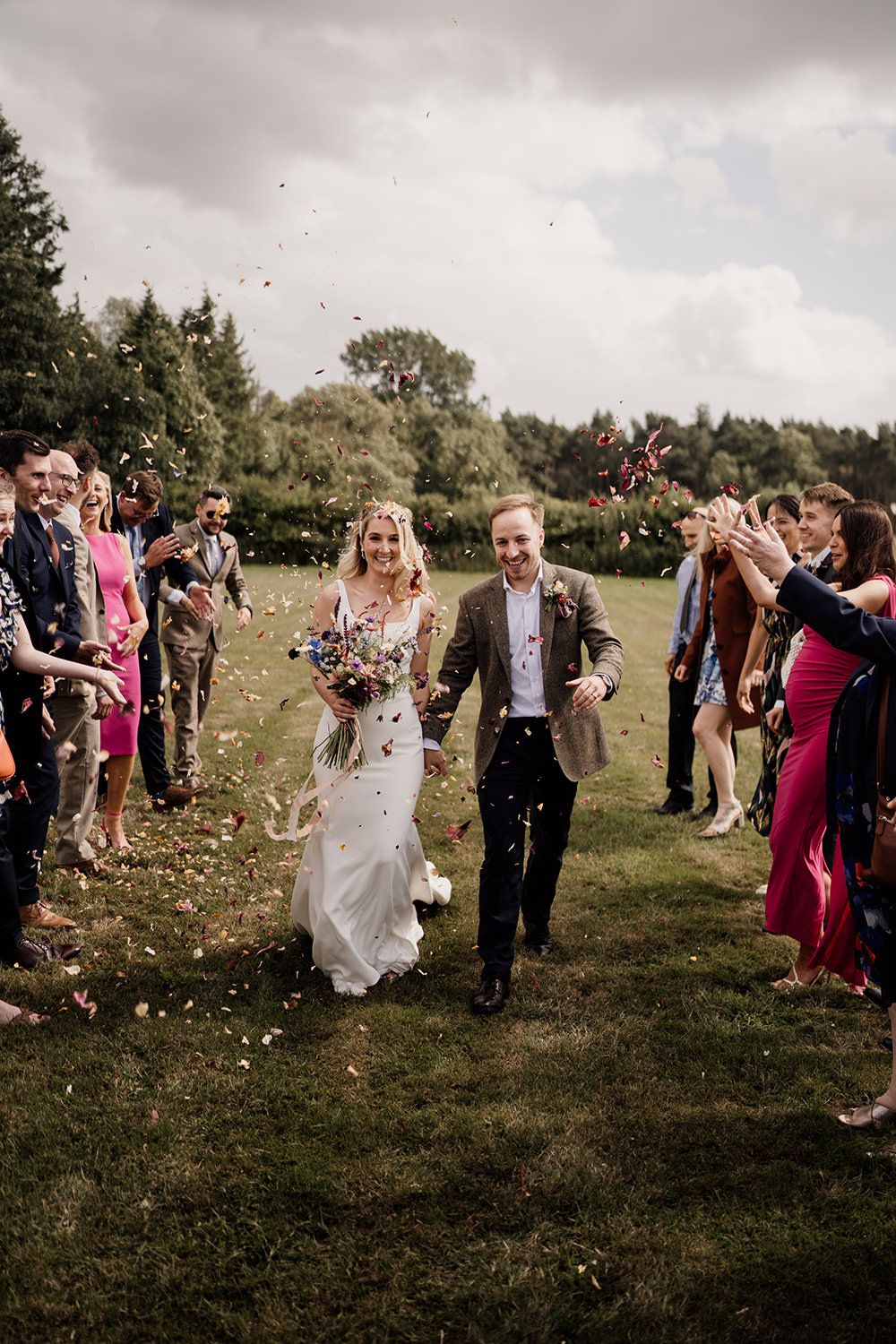 Bride and Groom holding hands smiling while guests form two lines and throw confetti in the grounds of Hovels Harewood.