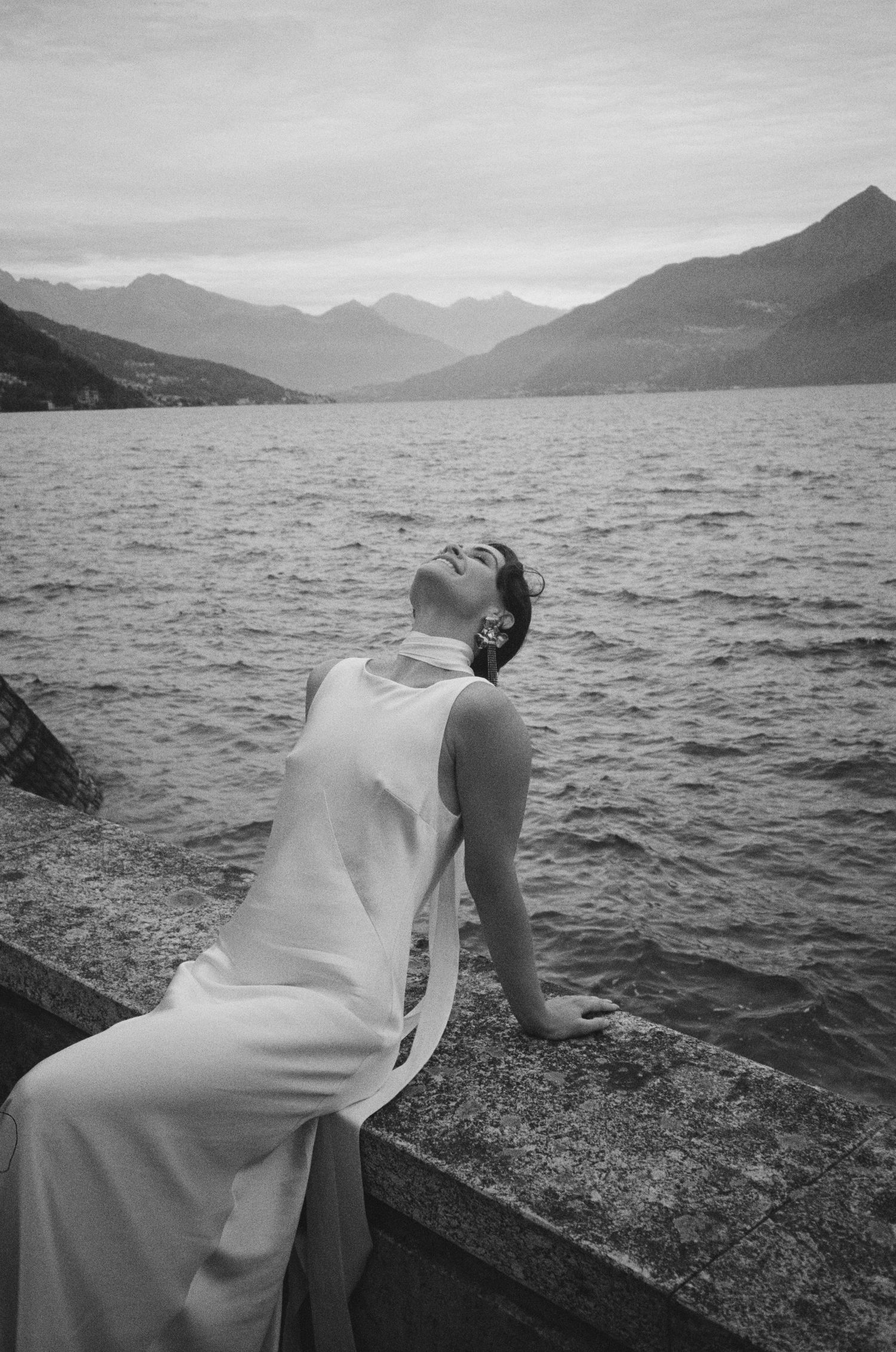 Black and white photo of a person in a white dress sitting by a mountain lake with dramatic peaks in the distance.