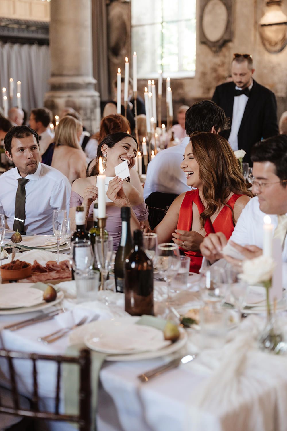 Wedding guests share laughter and conversation around elegant white-clothed dining tables at an indoor reception.