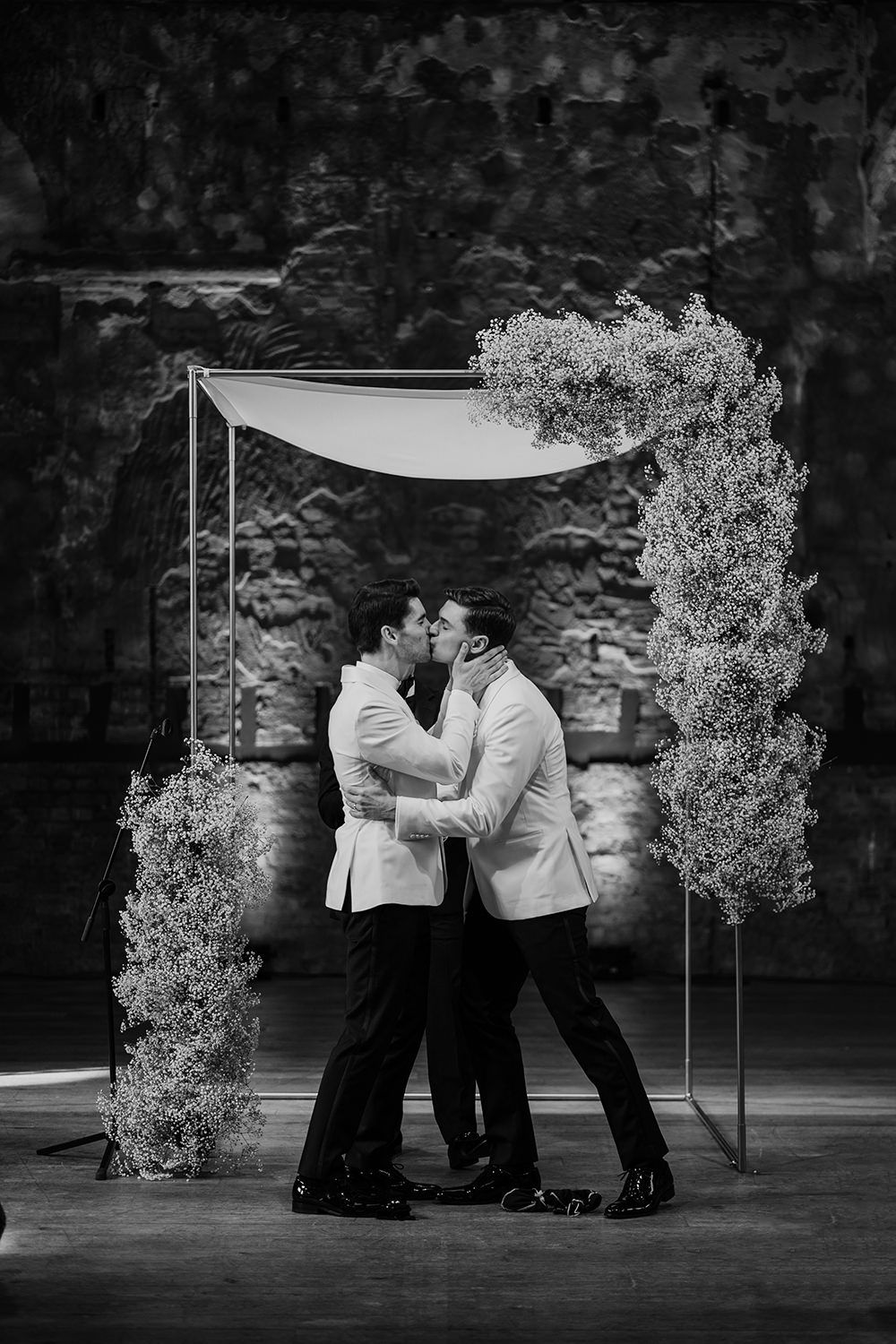 Two grooms and their first kiss, with cuppah and florals behind them.