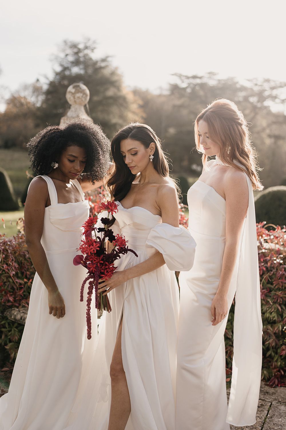 Three bridesmaids in elegant white wedding gowns pose outdoors, with one holding cascading burgundy flowers.