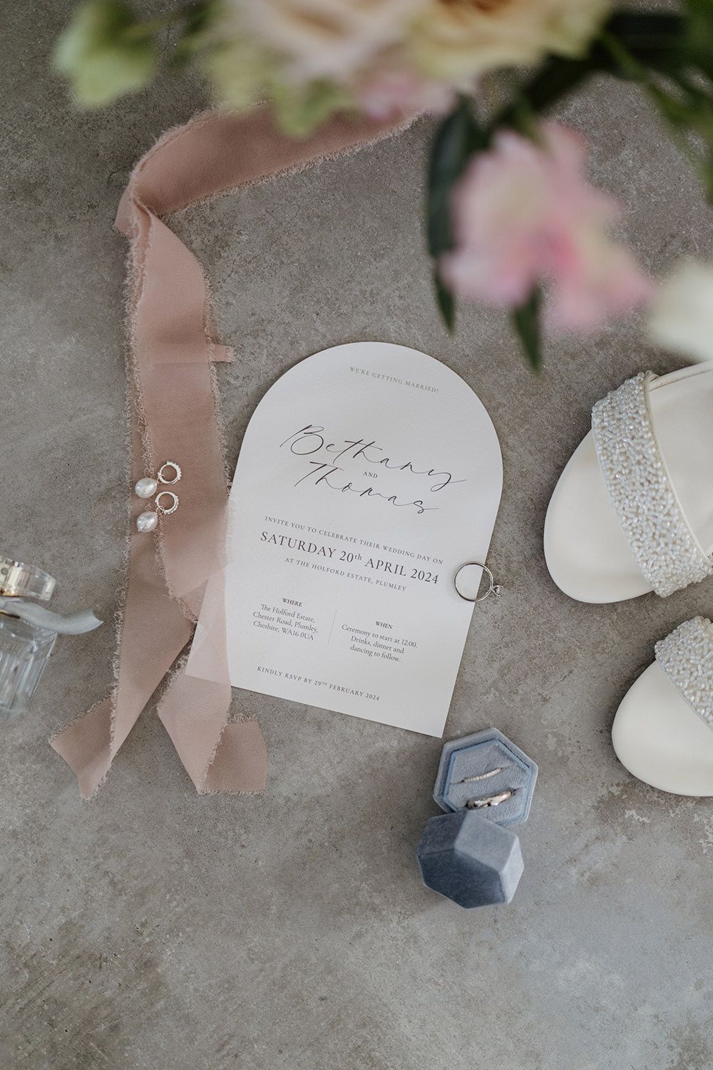 Wedding invite on grey floor with wedding rings, shoes, flowers and jewellery around.