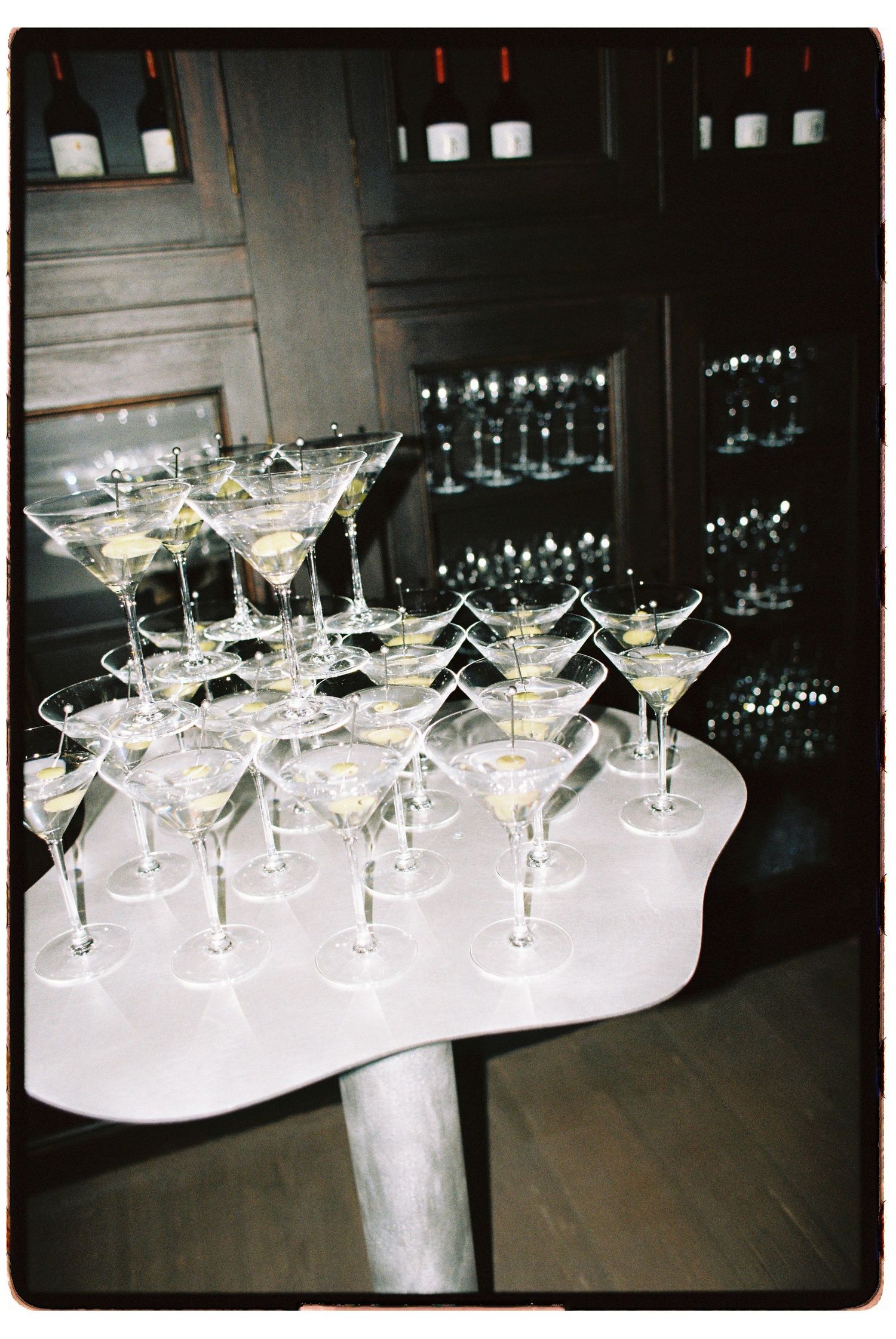 Elegant martini glasses arranged in a pyramid display on a white pedestal table at an upscale bar event.