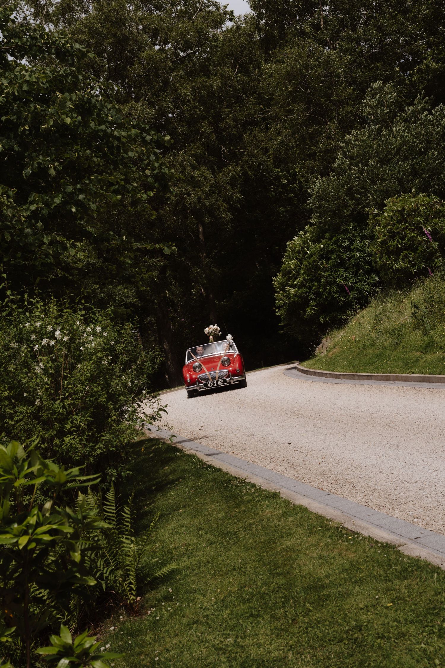 A vintage red convertible car drives down a scenic gravel road surrounded by lush green trees and manicured lawn.