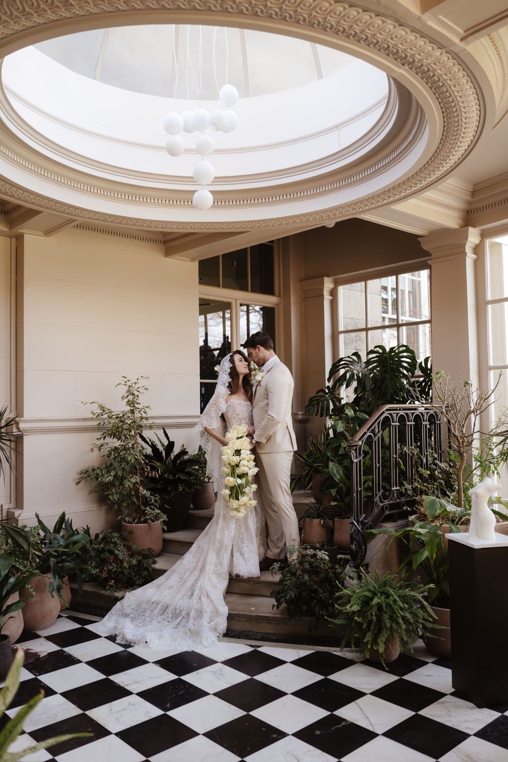 Bride and groom share a romantic moment in an elegant venue with checkered floors, lush greenery, and a stunning domed ceiling.