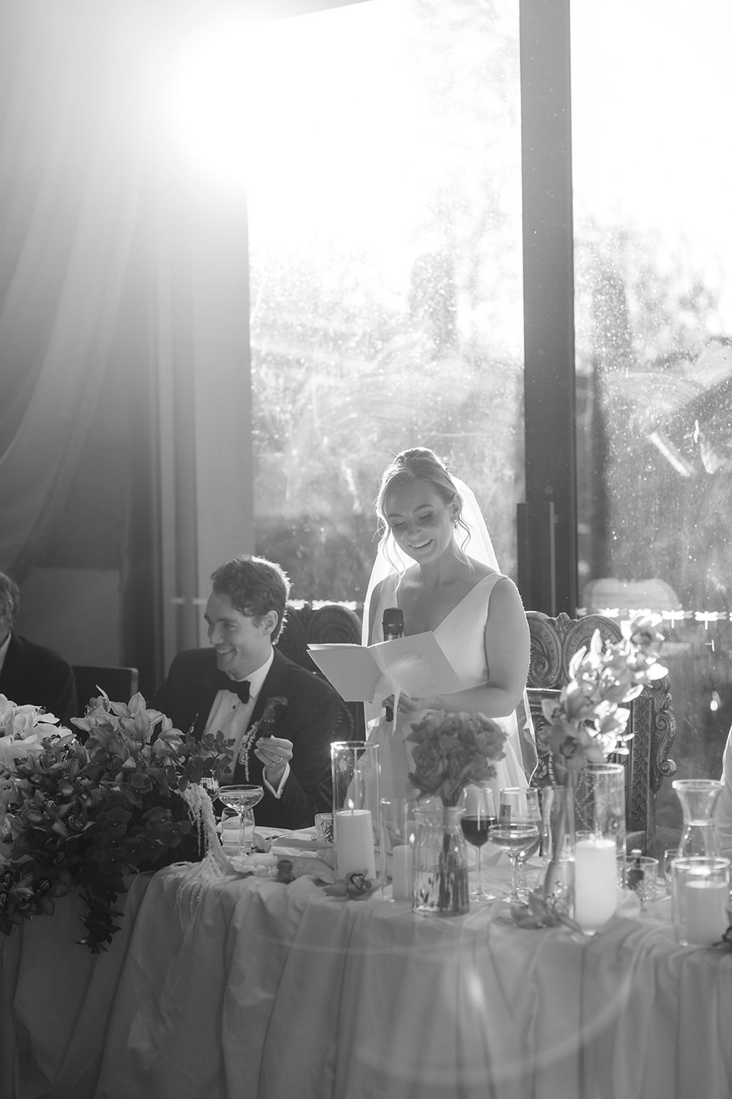 Bride smiles while reading vows at elegant wedding reception table with candles and floral arrangements in black and white photo.