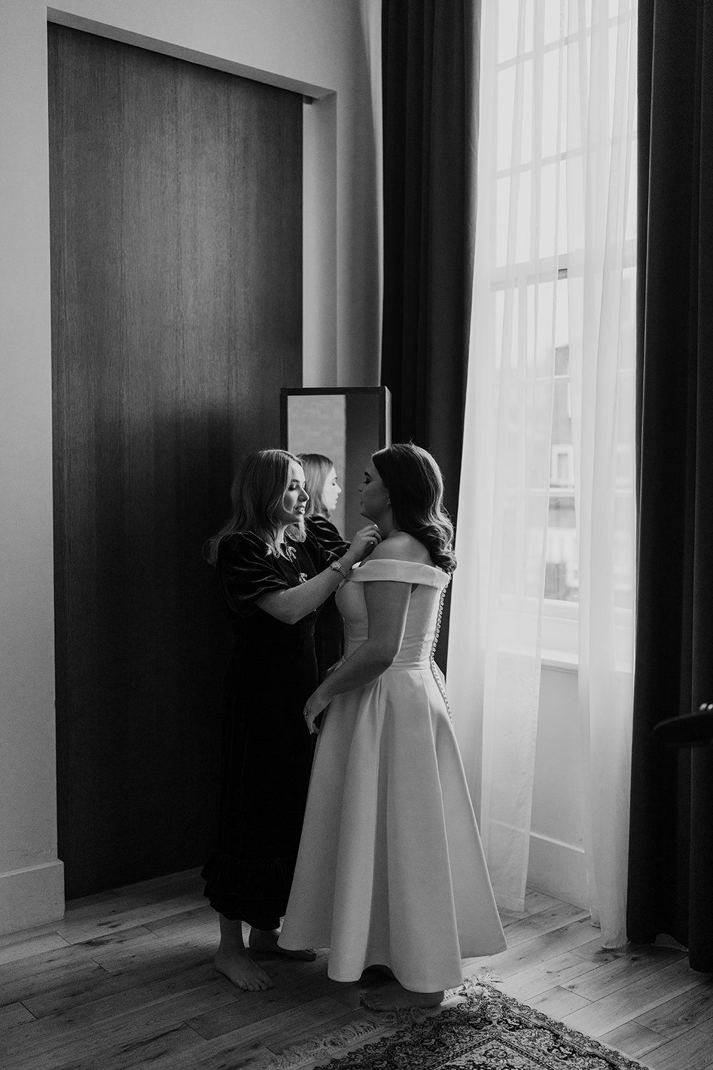 Black and white image of two people in a doorway, one wearing a white dress and another in dark clothing.