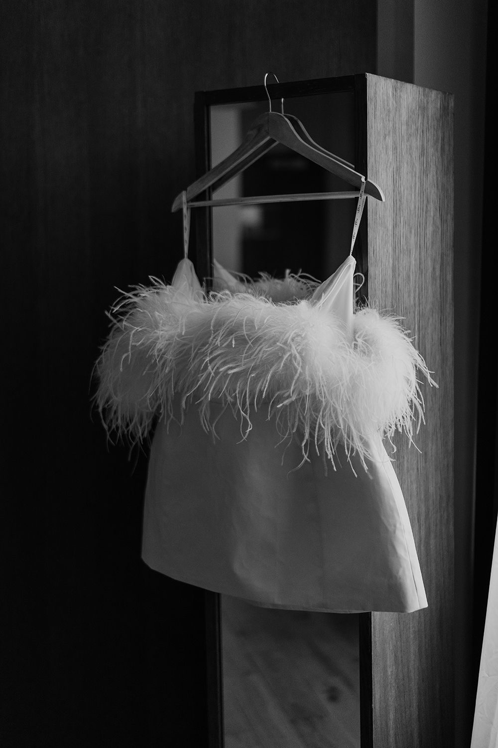 A white feather-trimmed dress hangs on a wooden hanger in dramatic black and white lighting.