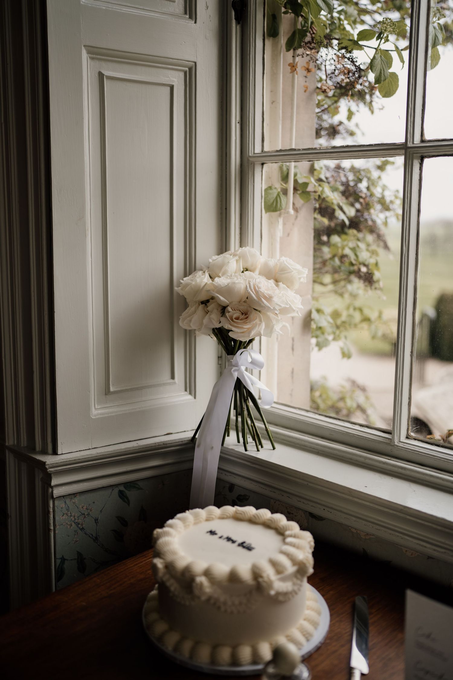 Wedding details in a window, long stem white roses and a white wedding cake.