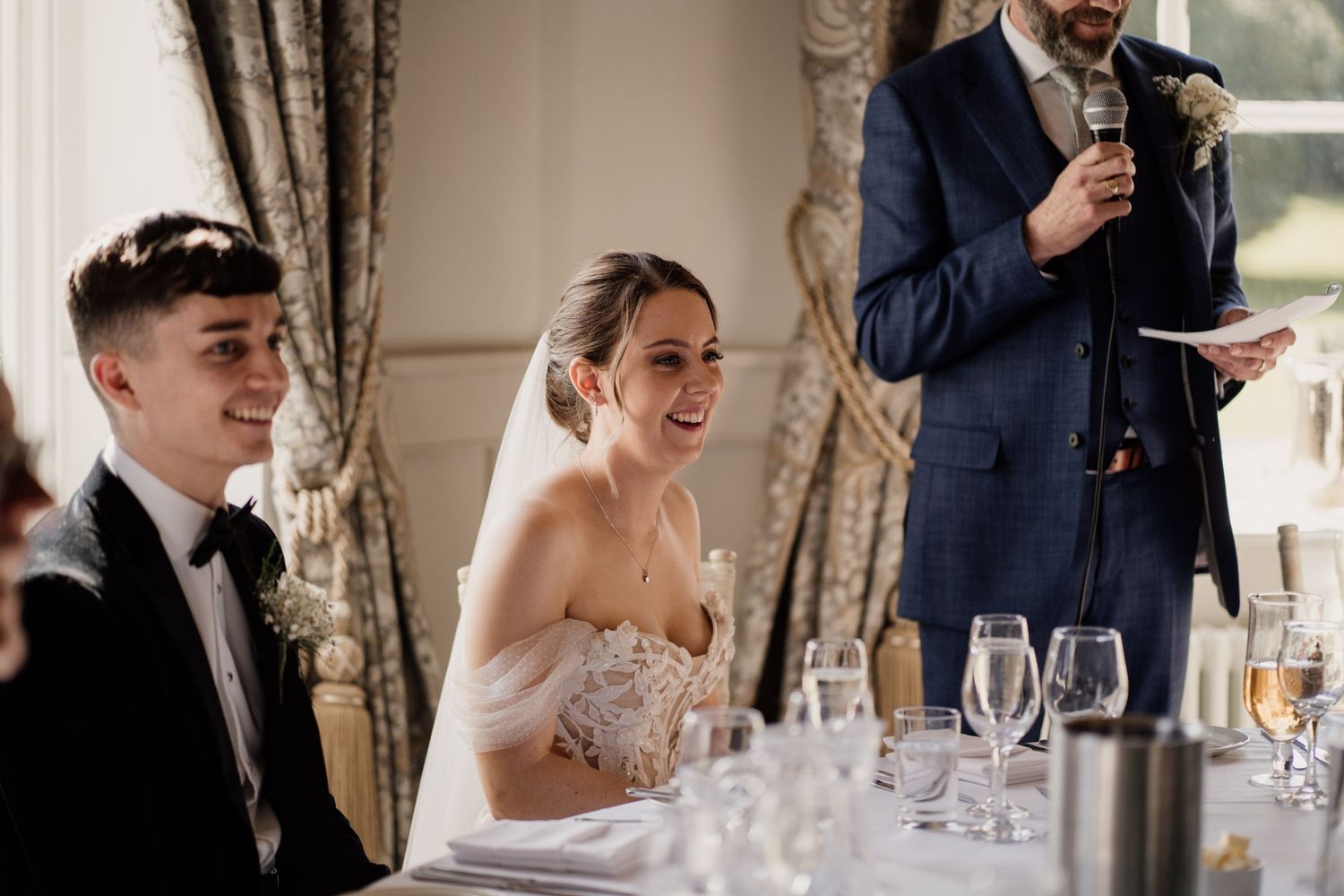 Bride smiling on her wedding day during speeches, sat at her reception table.