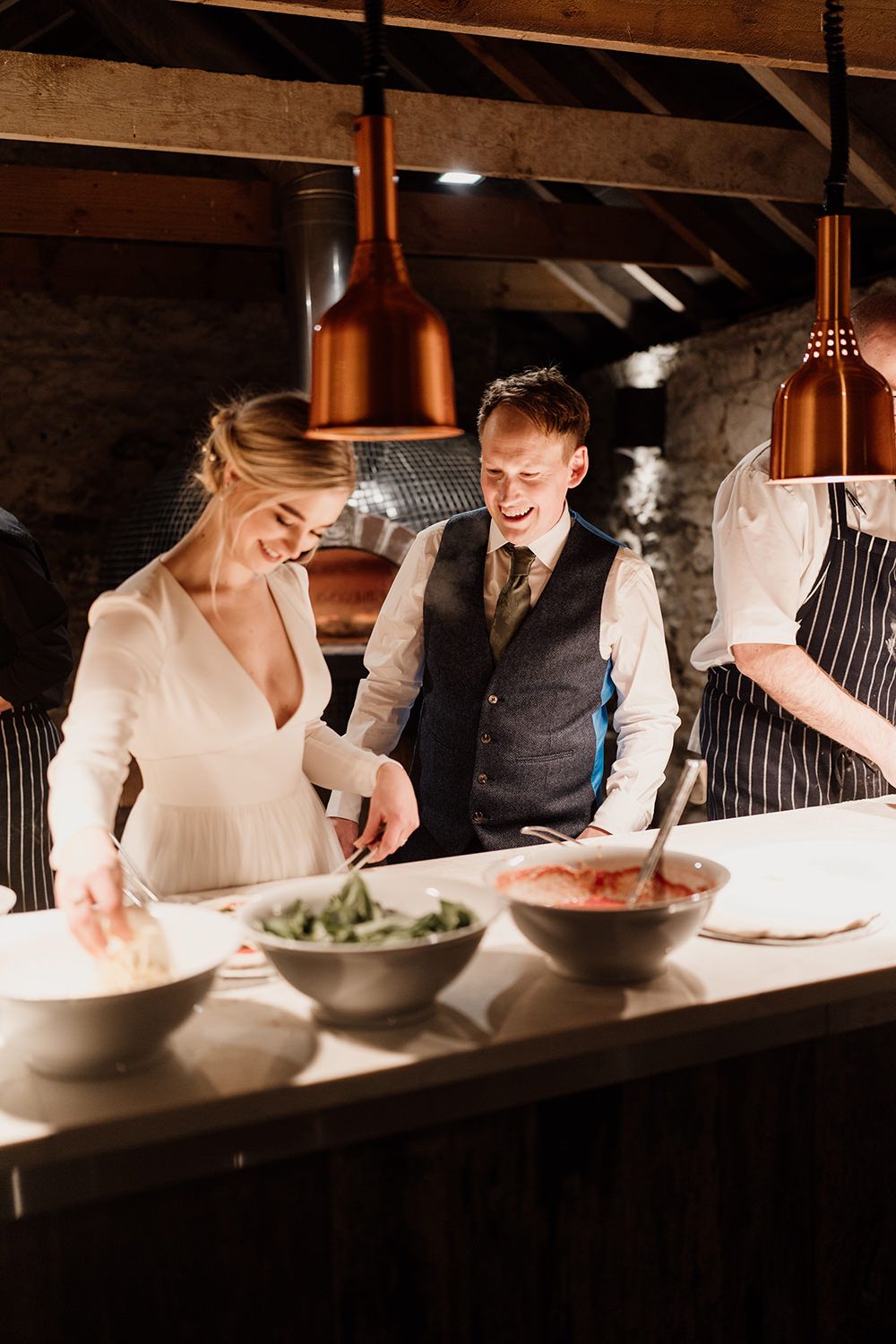 Bride and groom stood being shown how to make pizza on a white table.