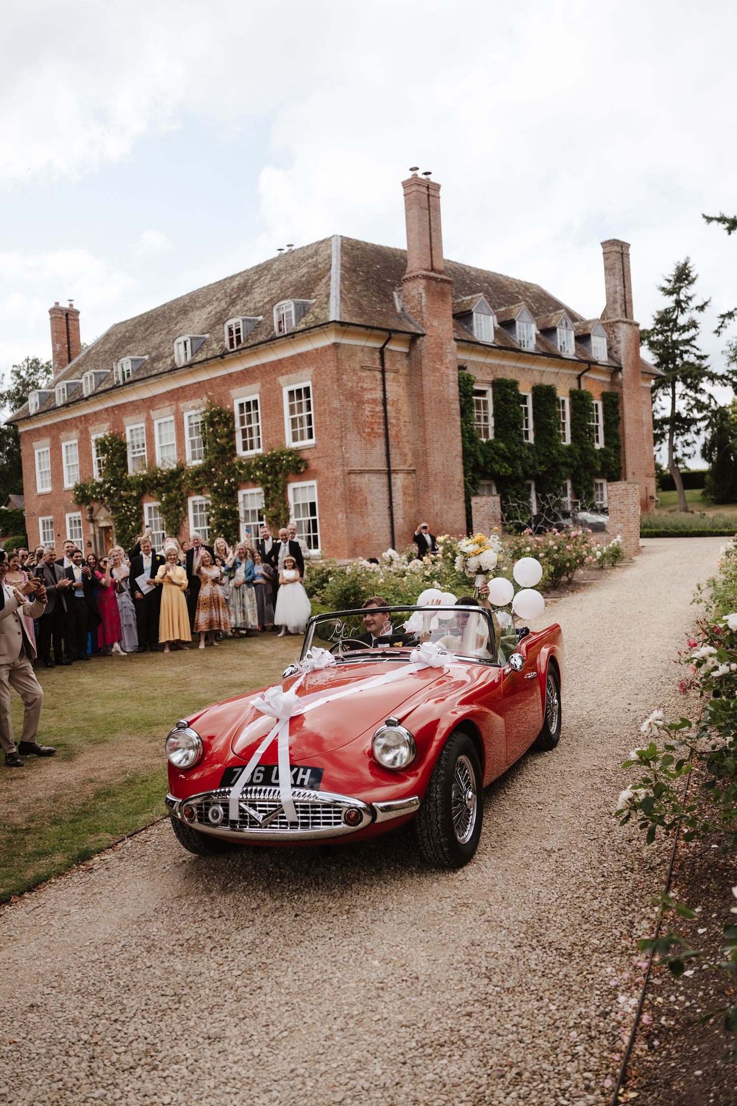 A vintage red convertible wedding car decorated with white ribbons drives past a grand brick manor house with guests cheering.