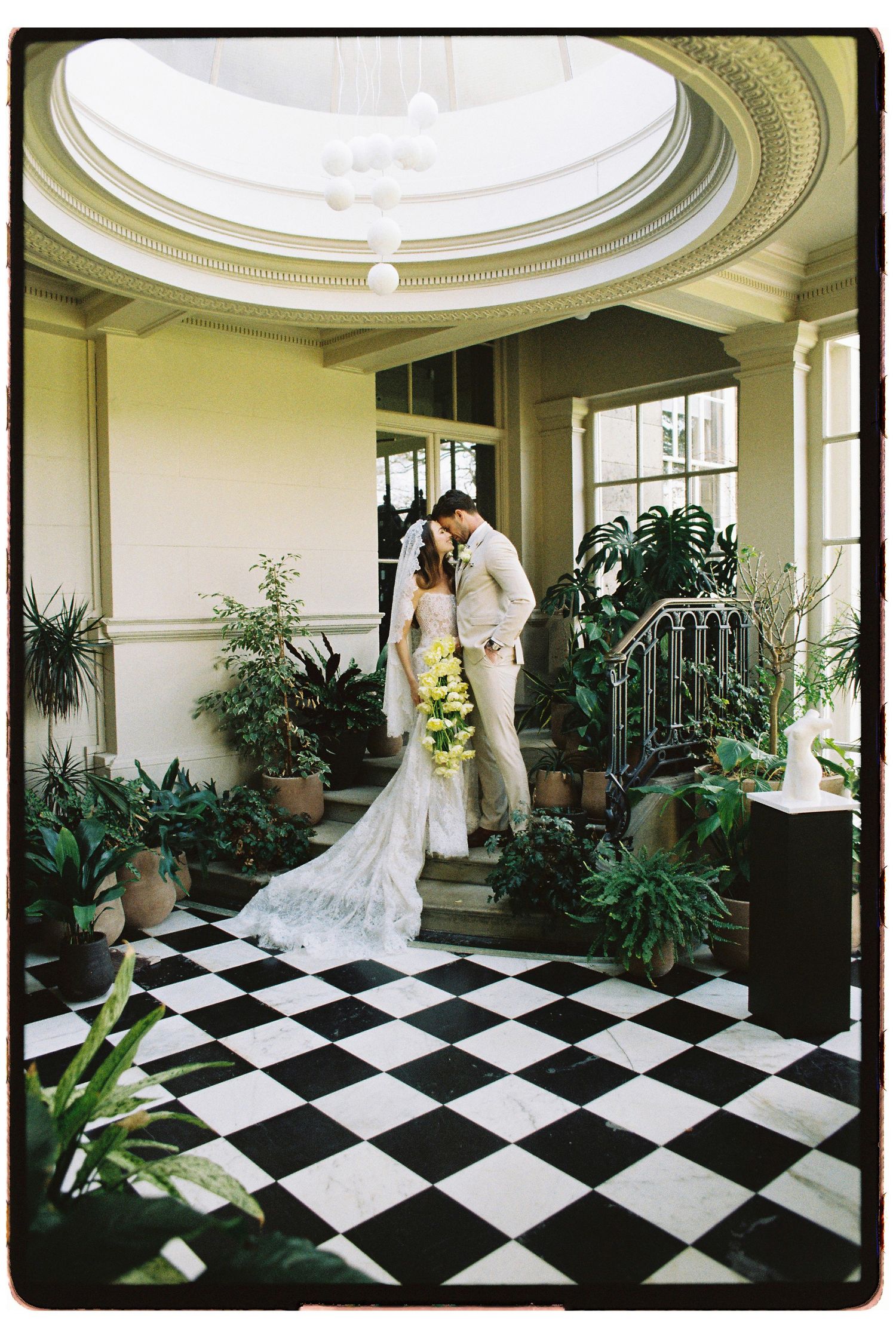 Bride and groom share a kiss in an elegant venue with black and white checkered floors and lush green plants.