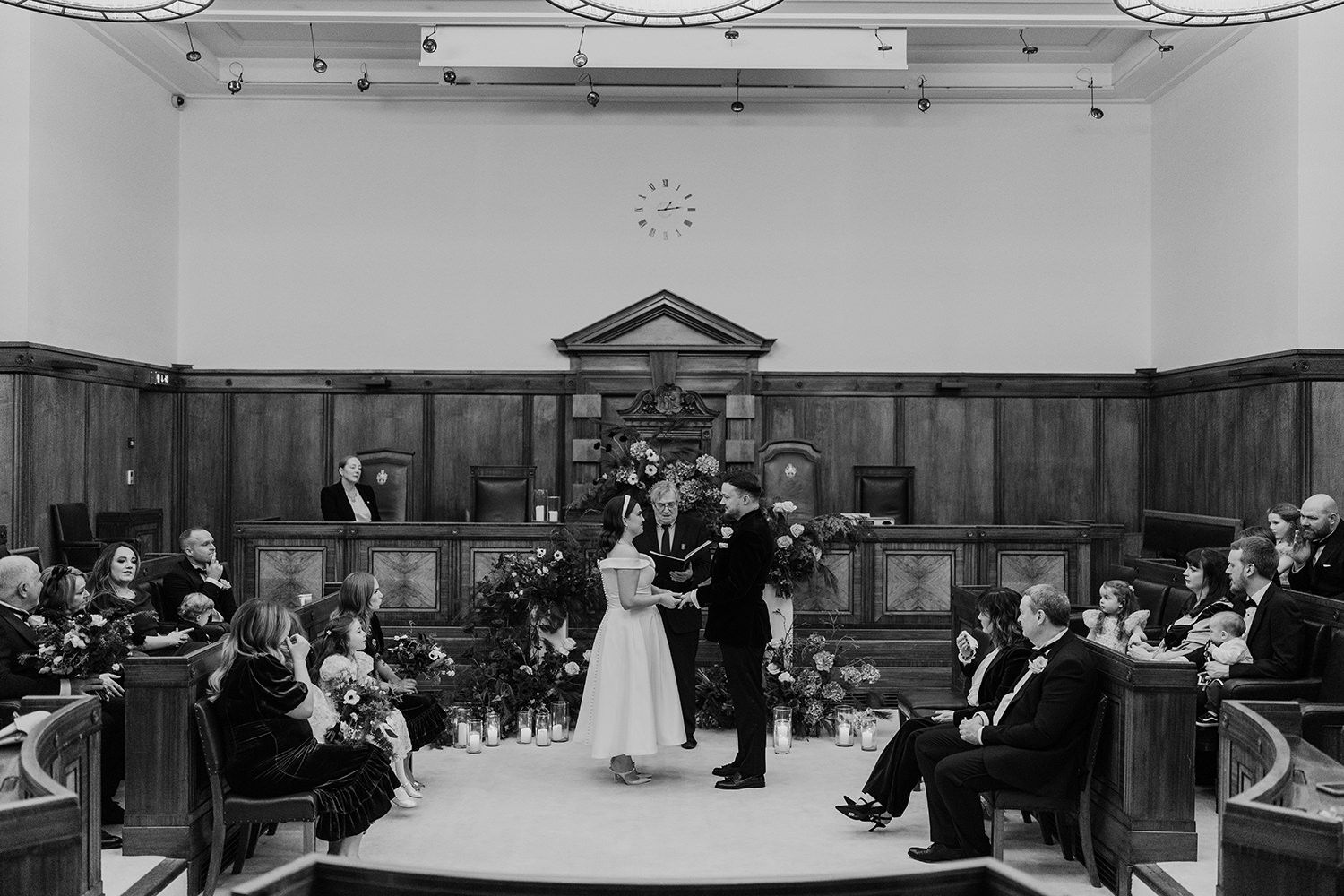 A black and white photograph of a wedding ceremony inside a historic courtroom with wooden paneling and gathered guests.