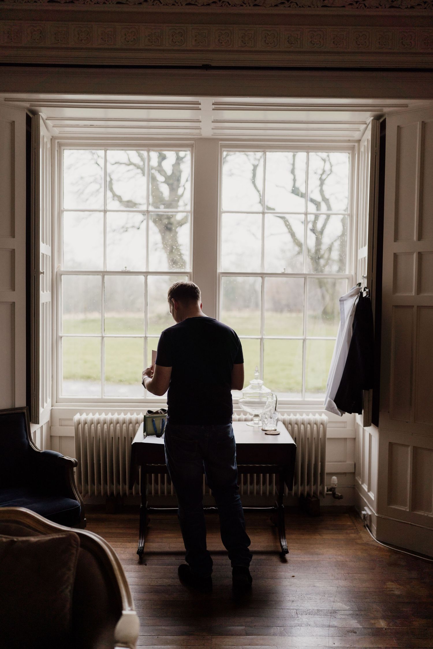 A person sits alone in dark silhouette against bright windows in an empty vintage room with radiators and wooden floors.