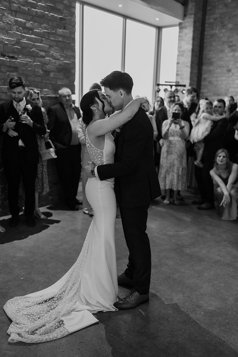 Bride and groom with arms around each other smiling as they do their first dance with friends and family watching.