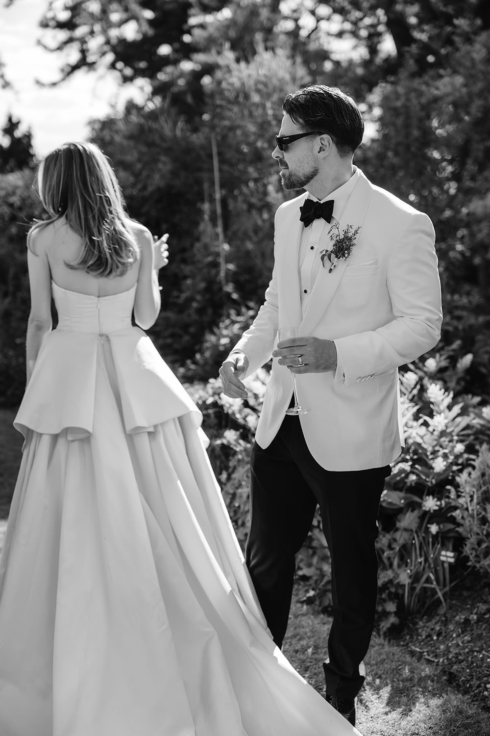 Black and white wedding photo of elegant couple in white dress and white tuxedo jacket outdoors among garden foliage.