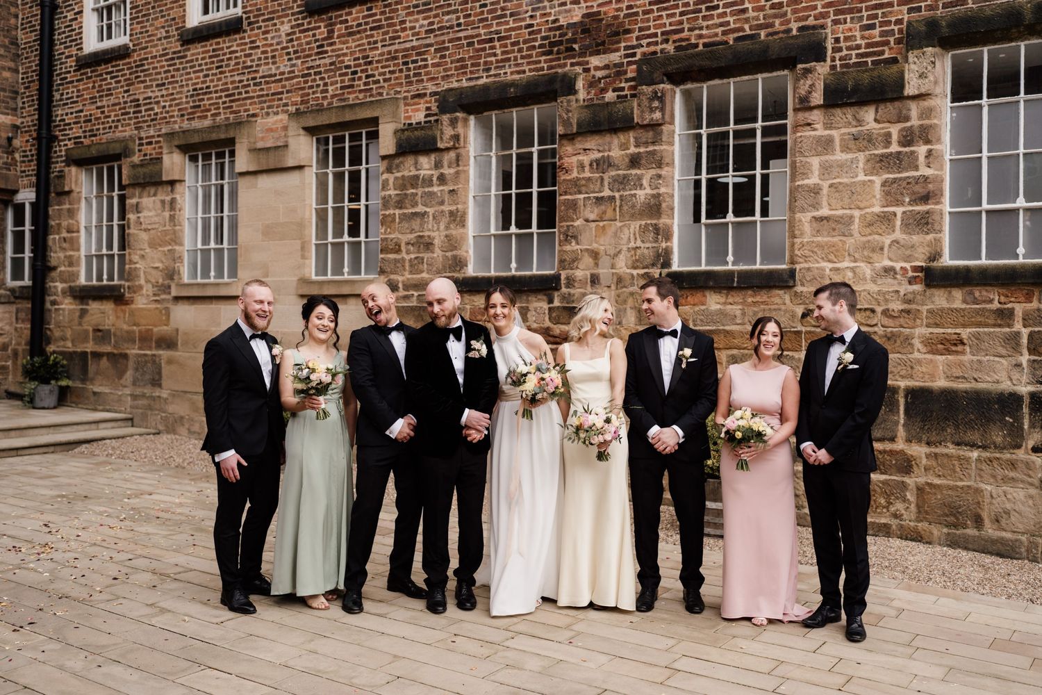 A wedding party poses for formal photos against historic stone architecture.