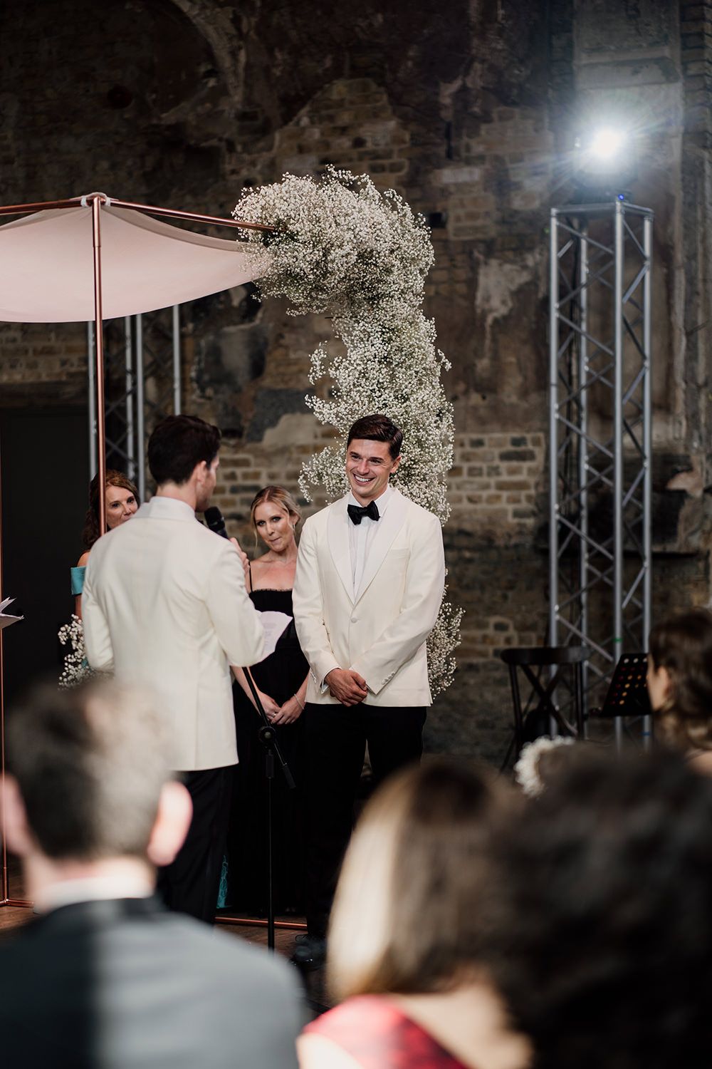 Grooms stood reading their wedding speeches, a white floral installation and studio lights are behind.