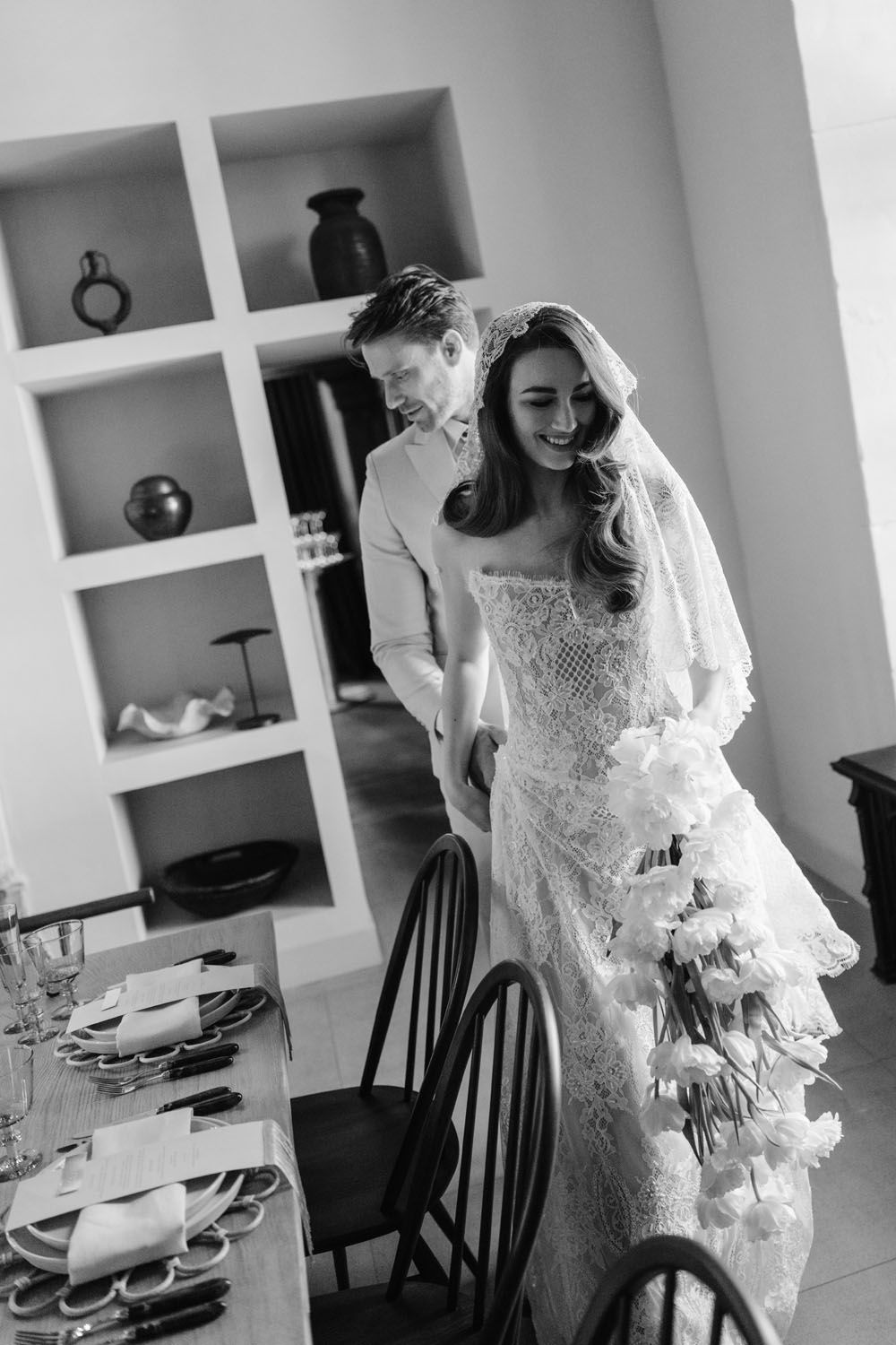 Black and white photo of a smiling bride in lace gown holding bouquet, walking with groom near elegantly set dining table.