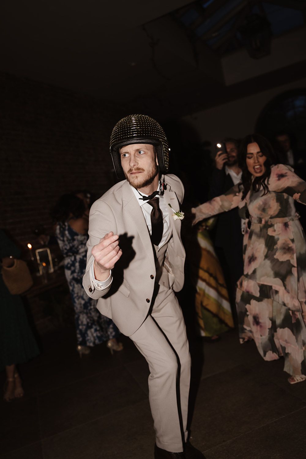 Person in light colored suit and helmet dances energetically at a party with others visible in background.