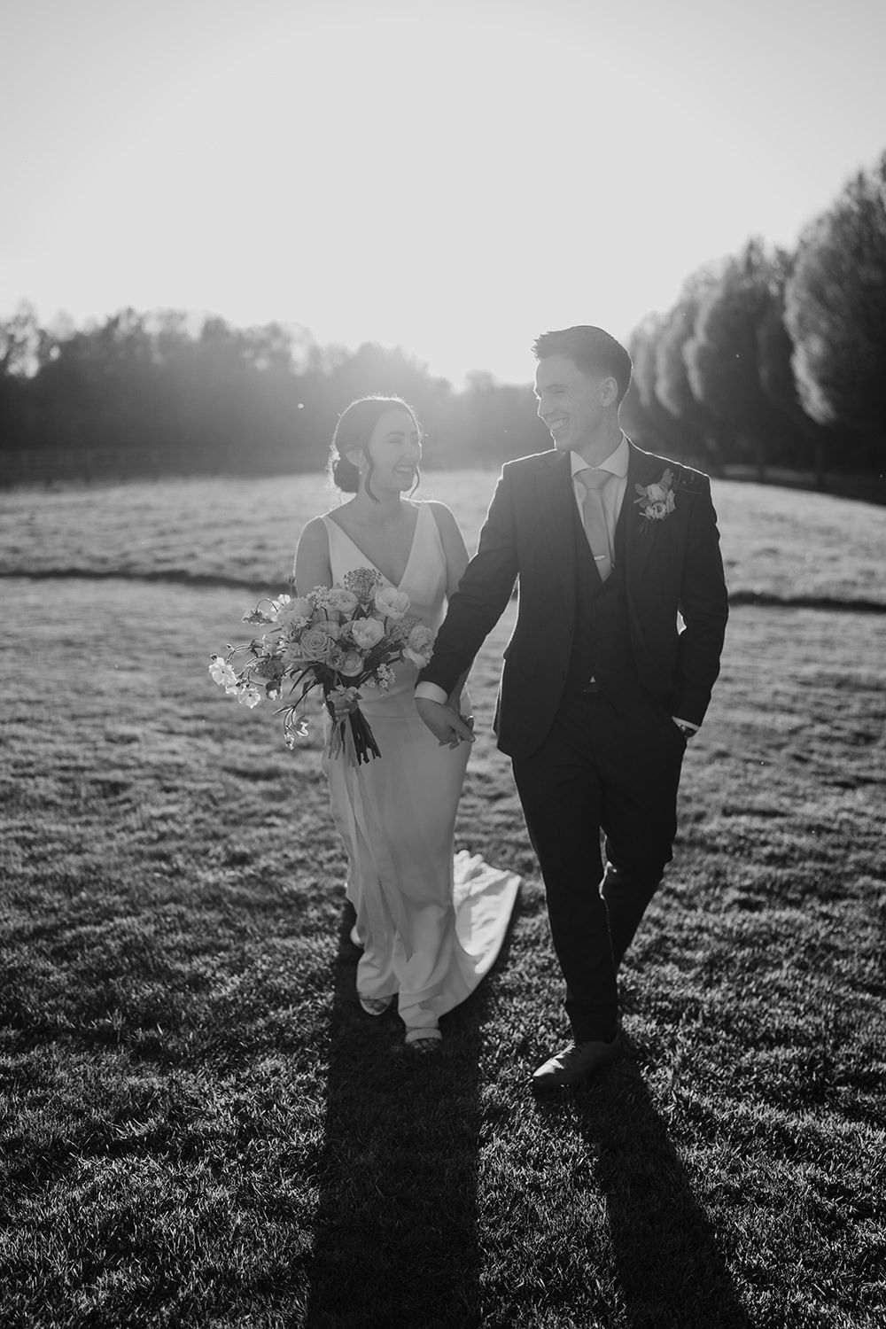Bride and groom walking across grass, laughing while holding hands with the sun setting.