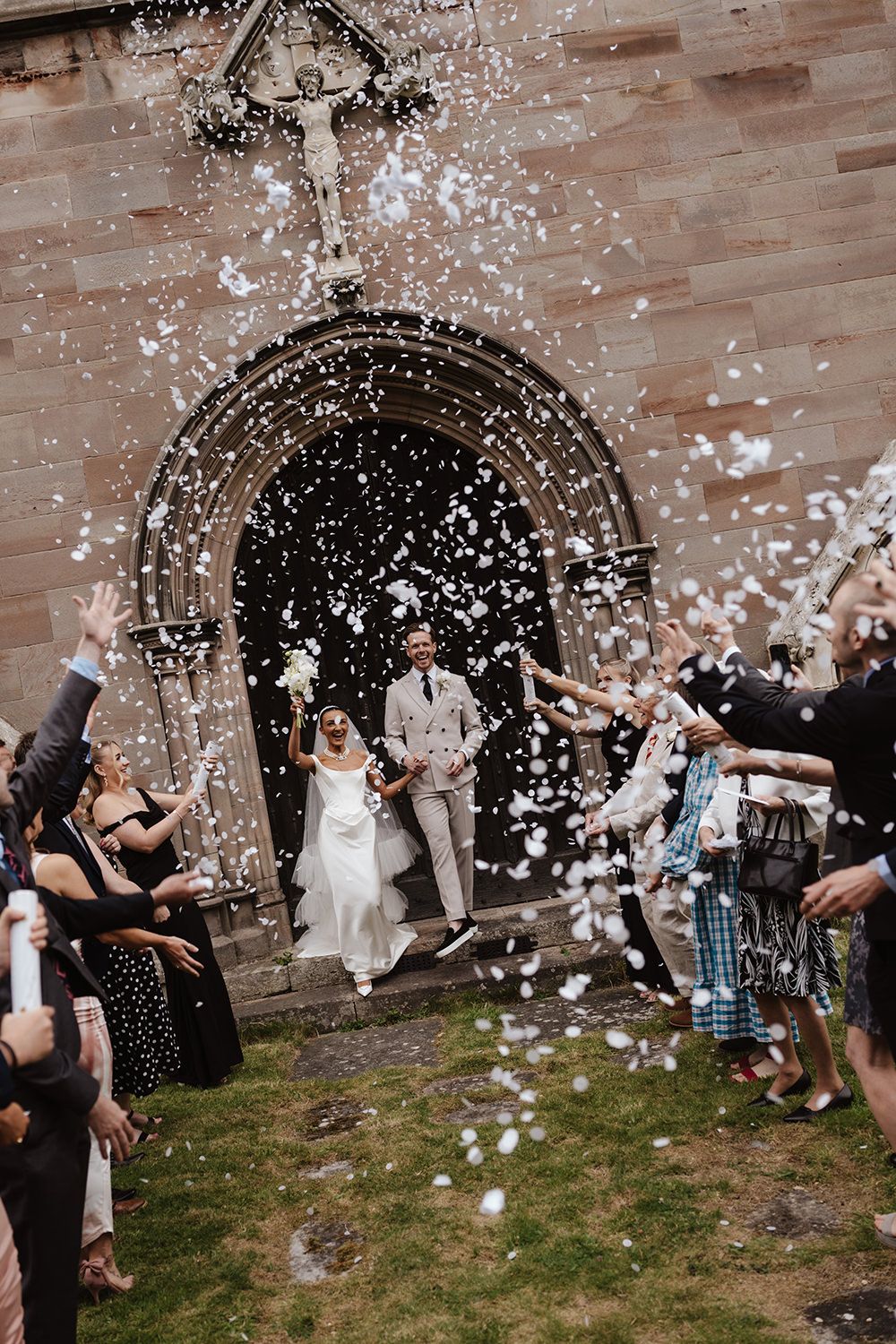 Wedding guests throw white confetti in celebration outside a stone archway at a joyous outdoor ceremony.