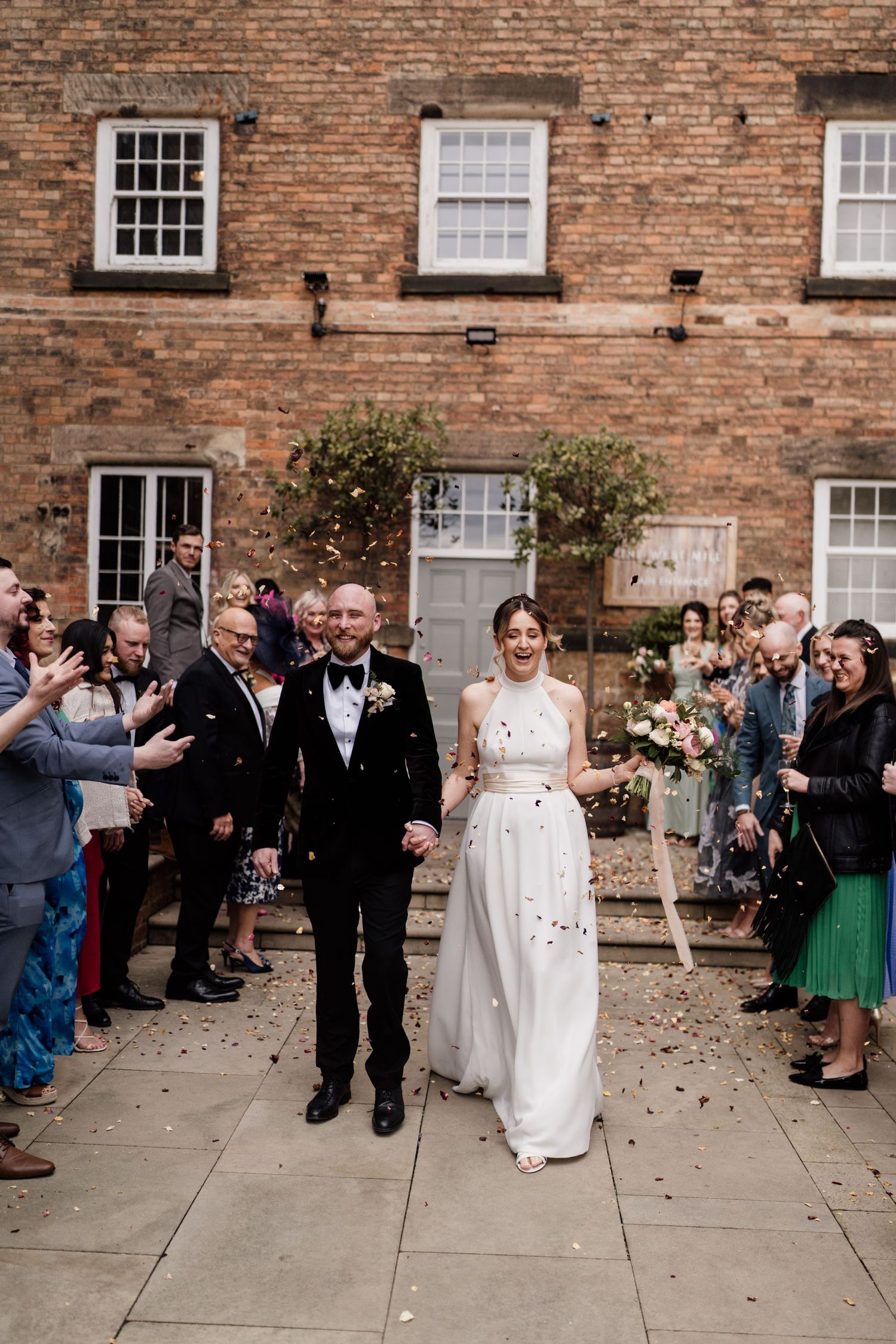 Wedding celebration outside historic brick building with guests throwing confetti.
