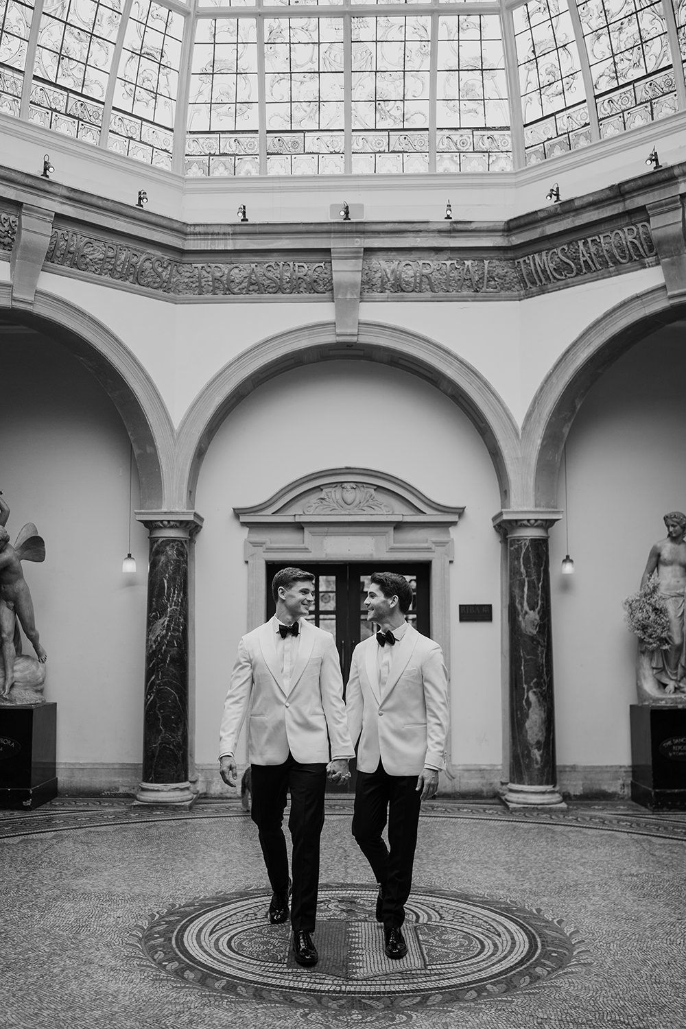 Two grooms holding hands walking towards camera while looking towards eachother, they're wearing monochrome suits.